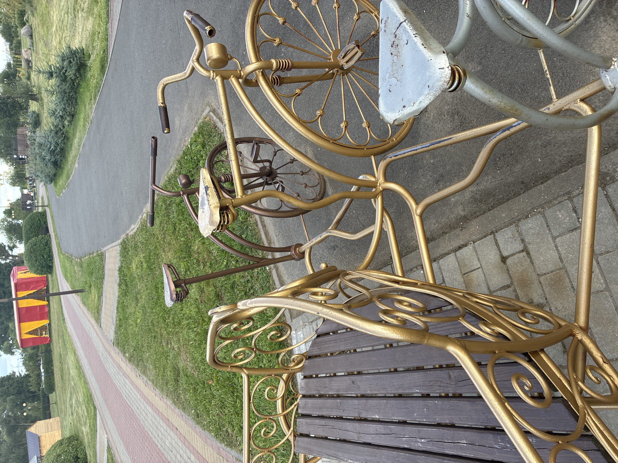 The image shows a decorative metal bench designed to resemble a tandem bicycle, placed outdoors on a paved area next to a grassy lawn. The bench is gold-colored and features ornate scrollwork. A wooden plank seat is visible on the bench. In the background, a paved pathway winds through a park-like setting. A red and yellow striped tent structure is visible in the distance, along with trees and some small buildings. The scene appears to be in a public park or recreational area in Mahilyow, Belarus. There are no people visible in the image.
