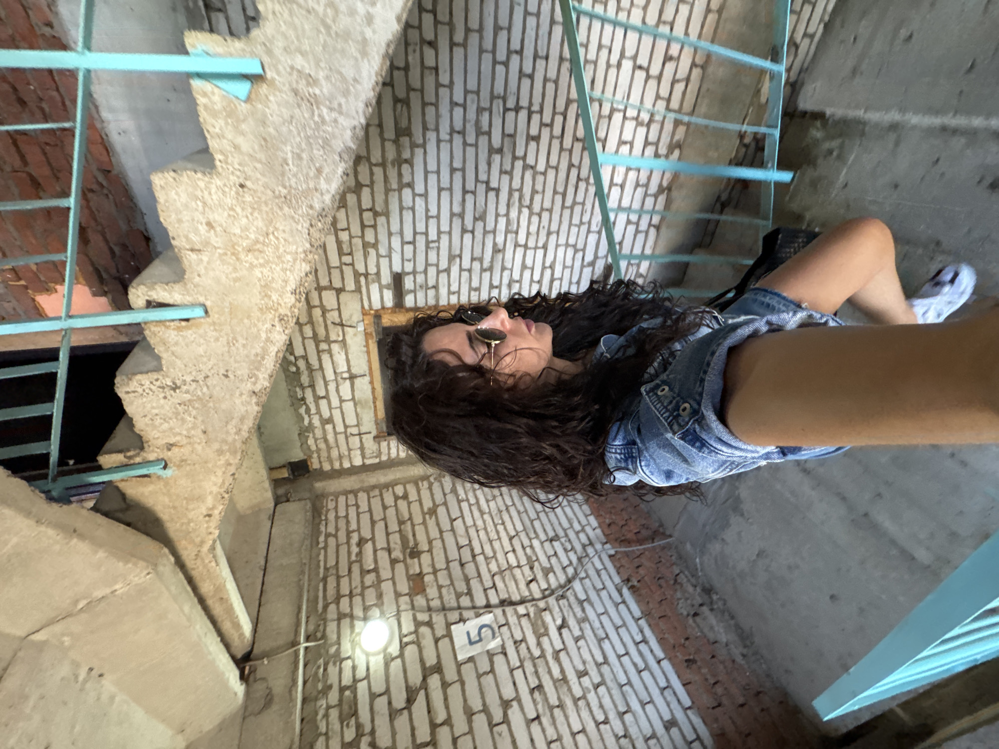A woman takes a selfie while standing in a stairwell. The walls of the stairwell are primarily white brick. The stair railing is a light blue color. The woman is wearing sunglasses and a denim jacket. A small sign with the number "5" is visible on the brick wall. The location is Chaikovsky, Russia.