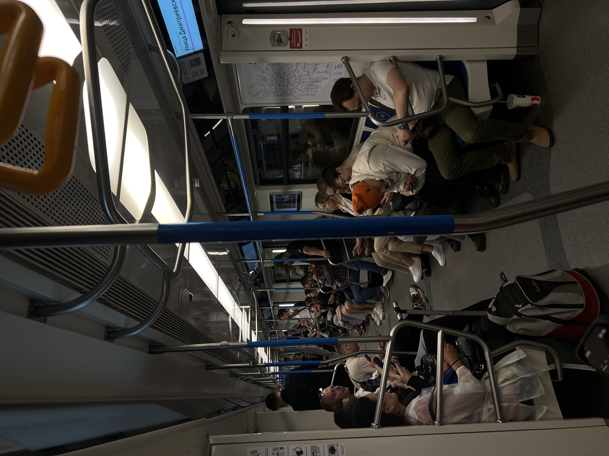 The image captures the interior of a crowded Moscow subway car. The passengers are seated, standing, and occupy the aisles; many are looking at or using mobile phones. The car's interior consists of white walls and ceilings, linear lighting, and blue handrails. A digital display shows the station name "Улица Дмитриевского" (Ulitsa Dmitrievskogo). A subway map is visible above the passengers. Personal belongings such as bags and suitcases are placed on the floor.