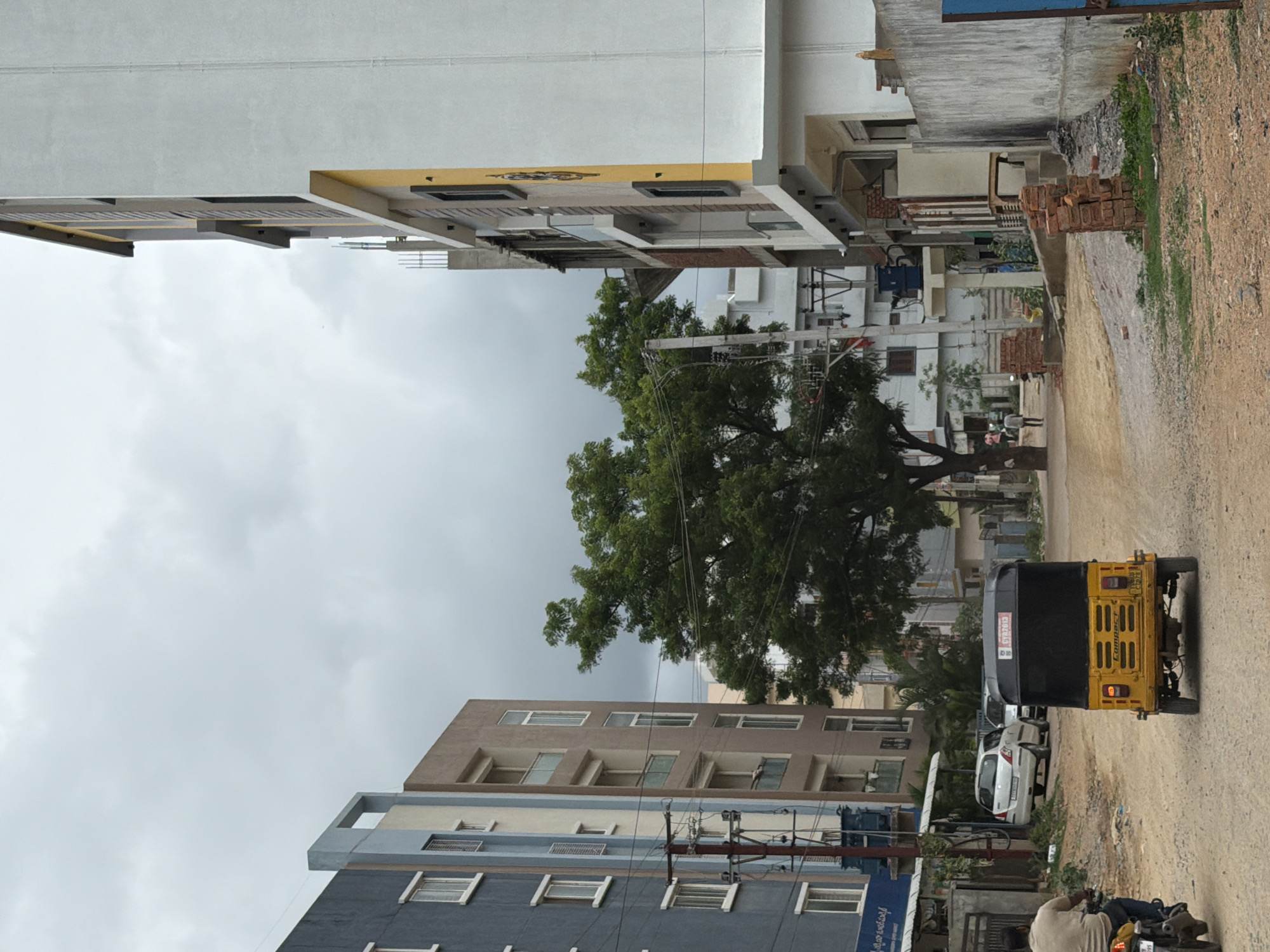 An overcast, daytime shot captures a street scene in Shamshabad, India, featuring a mix of residential and commercial buildings, vehicular traffic, and pedestrian activity. On the left, a blue and gray multi-story building stands next to a beige and white building. A power pole with multiple wires is situated in front of these structures. To the right, a prominent white and yellow building, also multi-story, is under construction or recently completed, with exposed brickwork and scaffolding visible in some areas. A large, leafy green tree is centered in the mid-ground, partially obscuring a smaller white building behind it. A yellow auto-rickshaw is parked on the dirt road in the foreground, facing away from the camera. To the left of the auto-rickshaw, a person on a motorcycle is moving away from the viewer. In the background, several cars are parked near a building, and a few pedestrians are walking on the right side of the street. The road surface is primarily dirt with some gravel and scattered bricks. The sky is cloudy and grey.