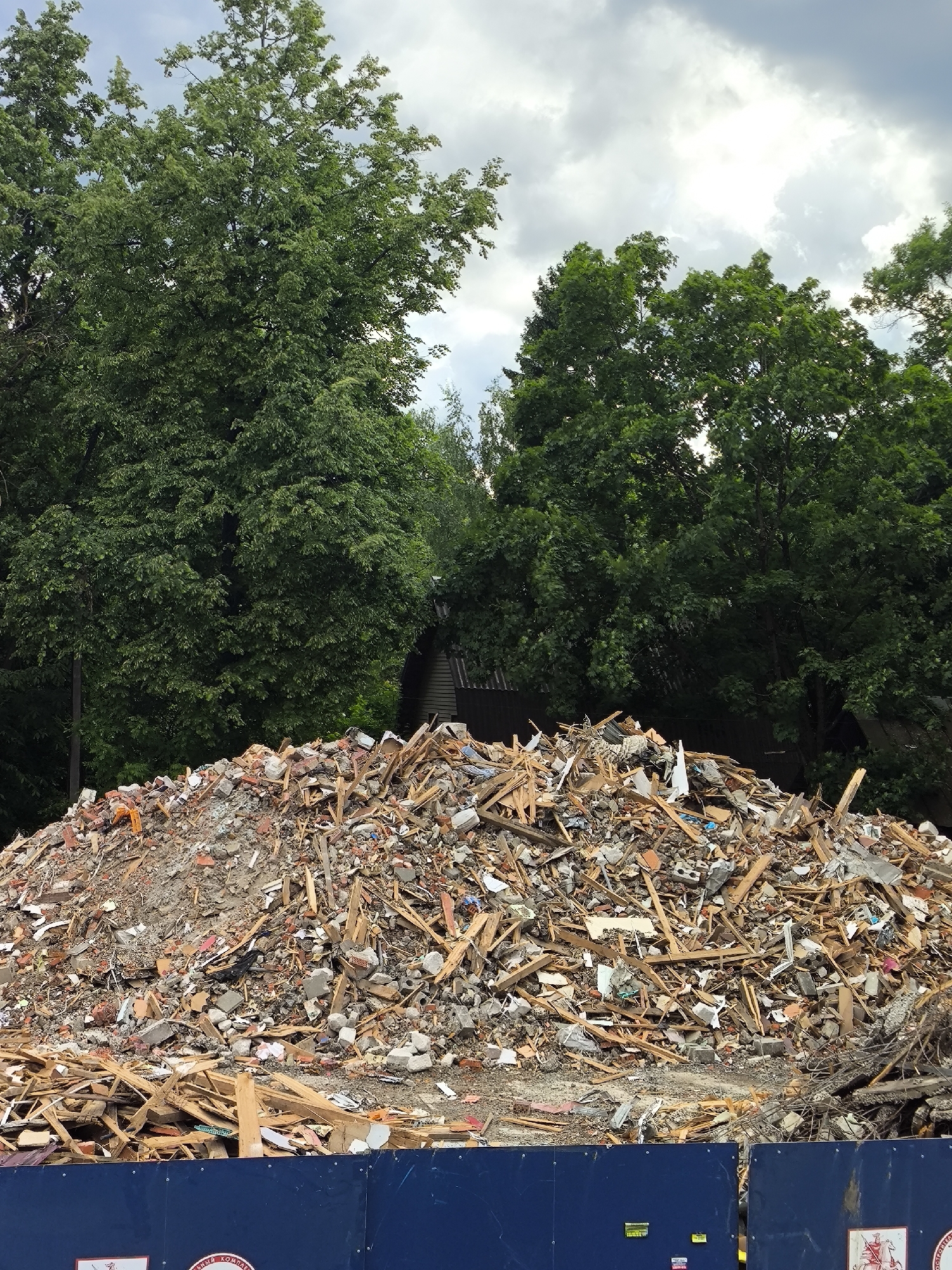 A large pile of debris consisting of broken bricks, wood scraps, concrete chunks, and metal fragments is situated in an outdoor environment. The pile is located in the city of Unknown, Unknown. A dark blue fence is positioned in front of the debris pile. Behind the debris pile, green trees and a structure are visible. The sky is cloudy.