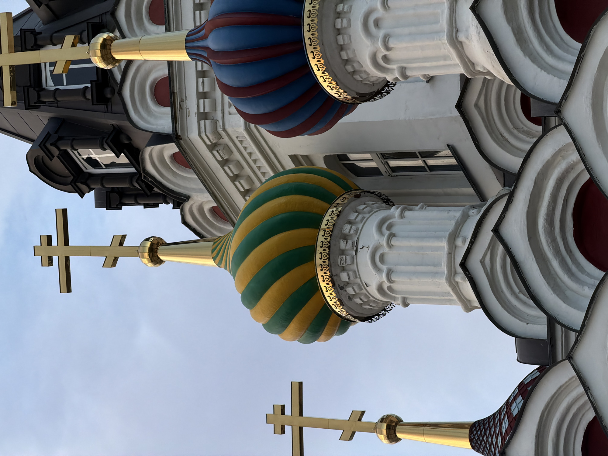 This is a close-up, low-angle shot of the domes and crosses of a church in Saratov, Russia. The sky is overcast with a pale blue hue. Three golden crosses, two of which are distinctly visible, crown the tops of the church's architectural elements. The central dome features a distinctive green and yellow striped pattern, while the dome to its right is adorned with alternating red and blue stripes. Both of these domes are topped with golden finials and elaborate golden ornamentation at their base. The church structure itself is predominantly white with white columns, architectural moldings, and arched window frames. The red bases of the domes are visible beneath the white detailing. The image captures a portion of the church's façade, emphasizing its ornate religious architecture.