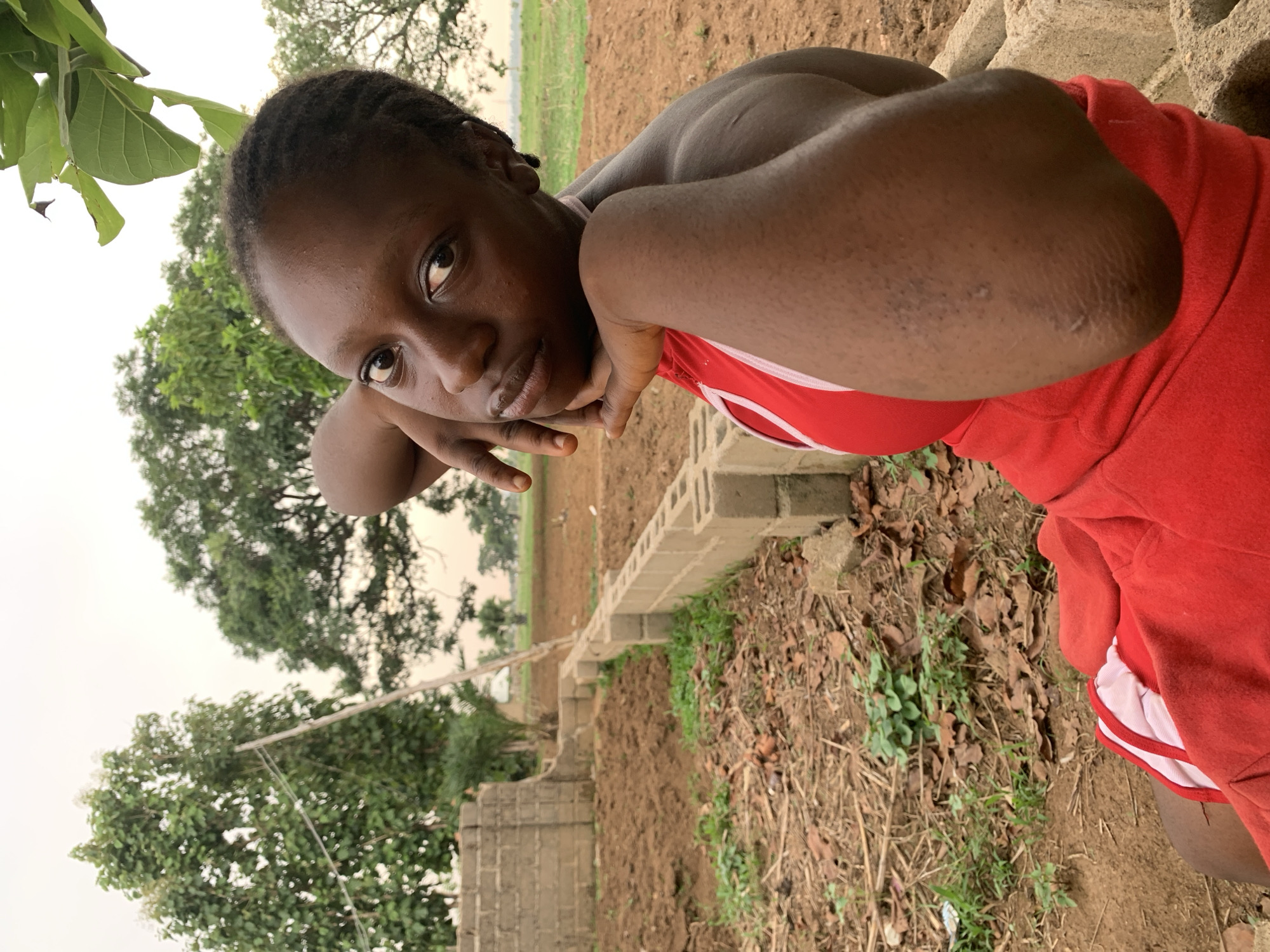 A young Black woman wearing a red top with white trim is positioned outdoors in Agban, Nigeria. She is leaning on her elbows, resting them on a structure made of grey concrete blocks. The woman's gaze is directed towards the camera. Behind her, a newly tilled patch of brown soil extends to the horizon, where a field of tall green grass is visible. To her left, a tree with green leaves is present, and further back, more trees are visible against a grey sky. The ground near the concrete structure is covered with dry leaves and some green vegetation. The building materials and recent digging suggest an active construction or agricultural site.