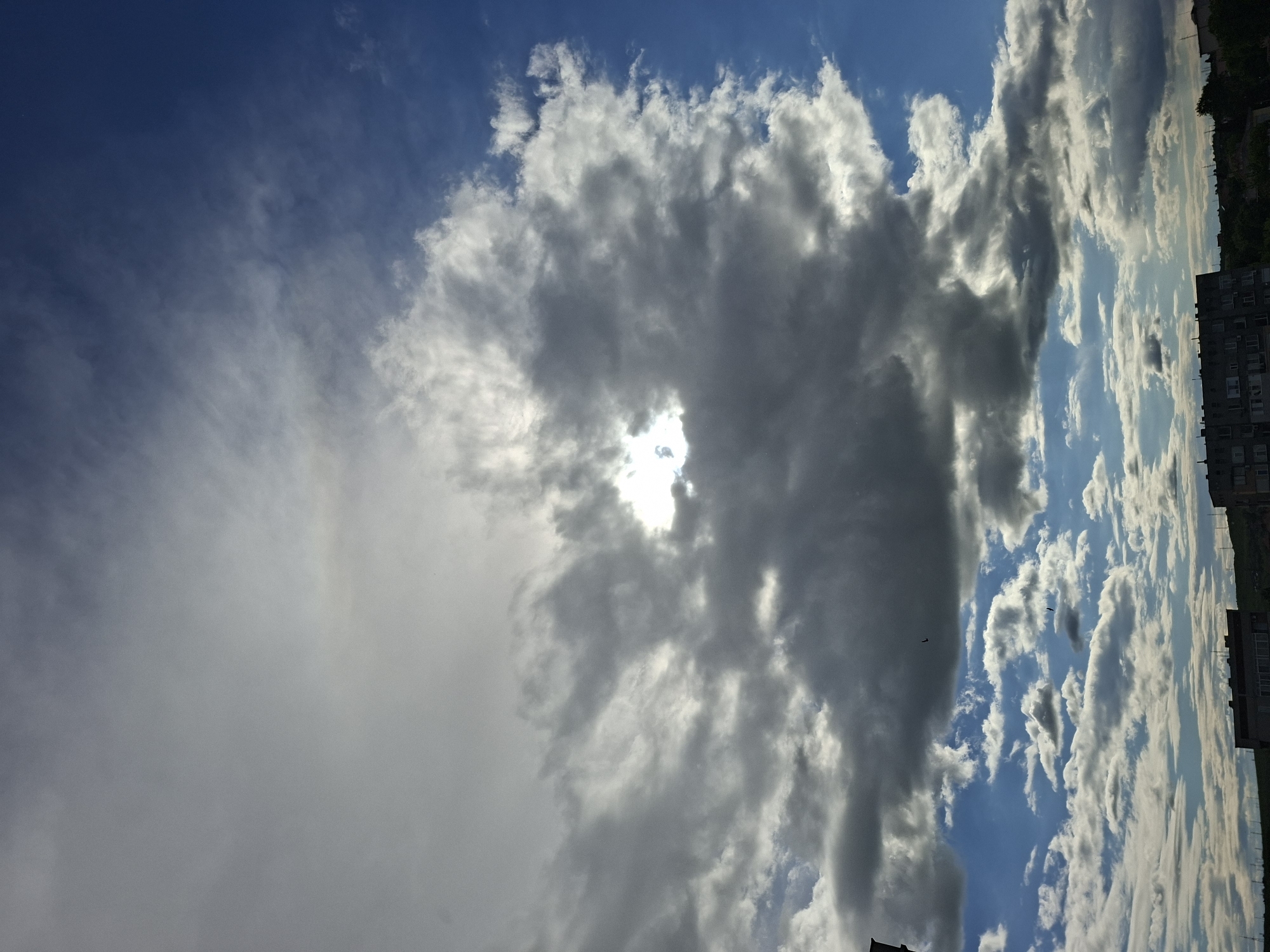 The image presents a sky with a mix of blue and clouds. A large, dark cloud dominates the central part of the frame, with a bright spot of sunlight peeking through it. To the left, a hazy, lighter cloud stretches across the sky. On the right side, there are more scattered, fluffy clouds against a blue backdrop. At the far right edge of the image, the tops of buildings are visible, suggesting an urban location, identified as Galabovo, Bulgaria. There's no clear indication of people, actions, or specific interactions within the frame. The environment is primarily defined by the atmospheric conditions and the presence of city structures at the margin.