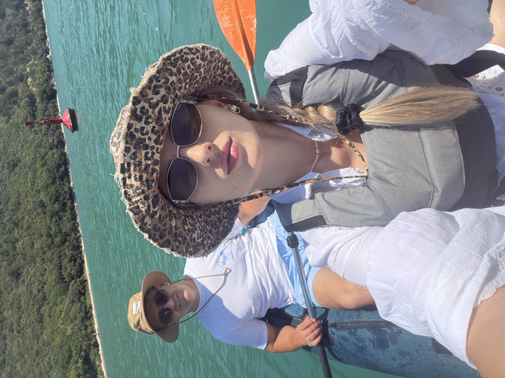 A man and a woman are kayaking on a body of water. The man is in the foreground, wearing a white t-shirt, blue shorts, sunglasses, and a tan bucket hat. He is holding a paddle and smiling at the camera. The woman is behind him, wearing a white shirt, a grey life vest, sunglasses, and a leopard-print bucket hat. She is also holding a paddle and looking at the camera. The water is a clear turquoise color, and there are green trees on the shore in the background. A red buoy is visible in the water in the distance. The scene appears to be a sunny day.