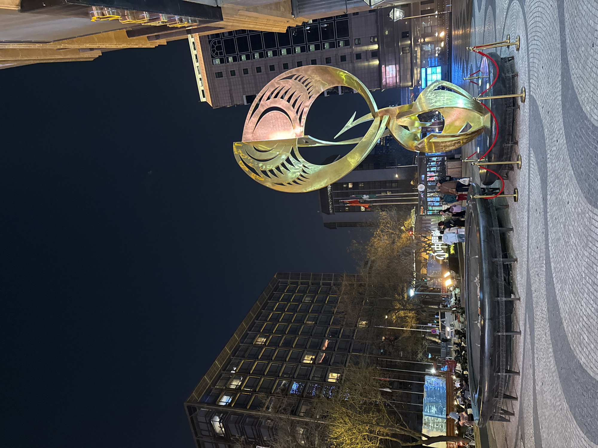 This nighttime photograph shows a large, golden metal sculpture in the foreground of a city square, likely in Dongcheng District, China. The sculpture appears to be abstract, possibly depicting stylized fish or leaves. It is surrounded by a rope barrier supported by gold posts. In the background, several multistory buildings with lighted windows are visible, indicating an urban setting. Some people are gathered further back in the square, near what seems to be a display or stage illuminated by bright lights. Bare tree branches are visible in the middle ground. The ground surface is covered in a patterned mosaic.