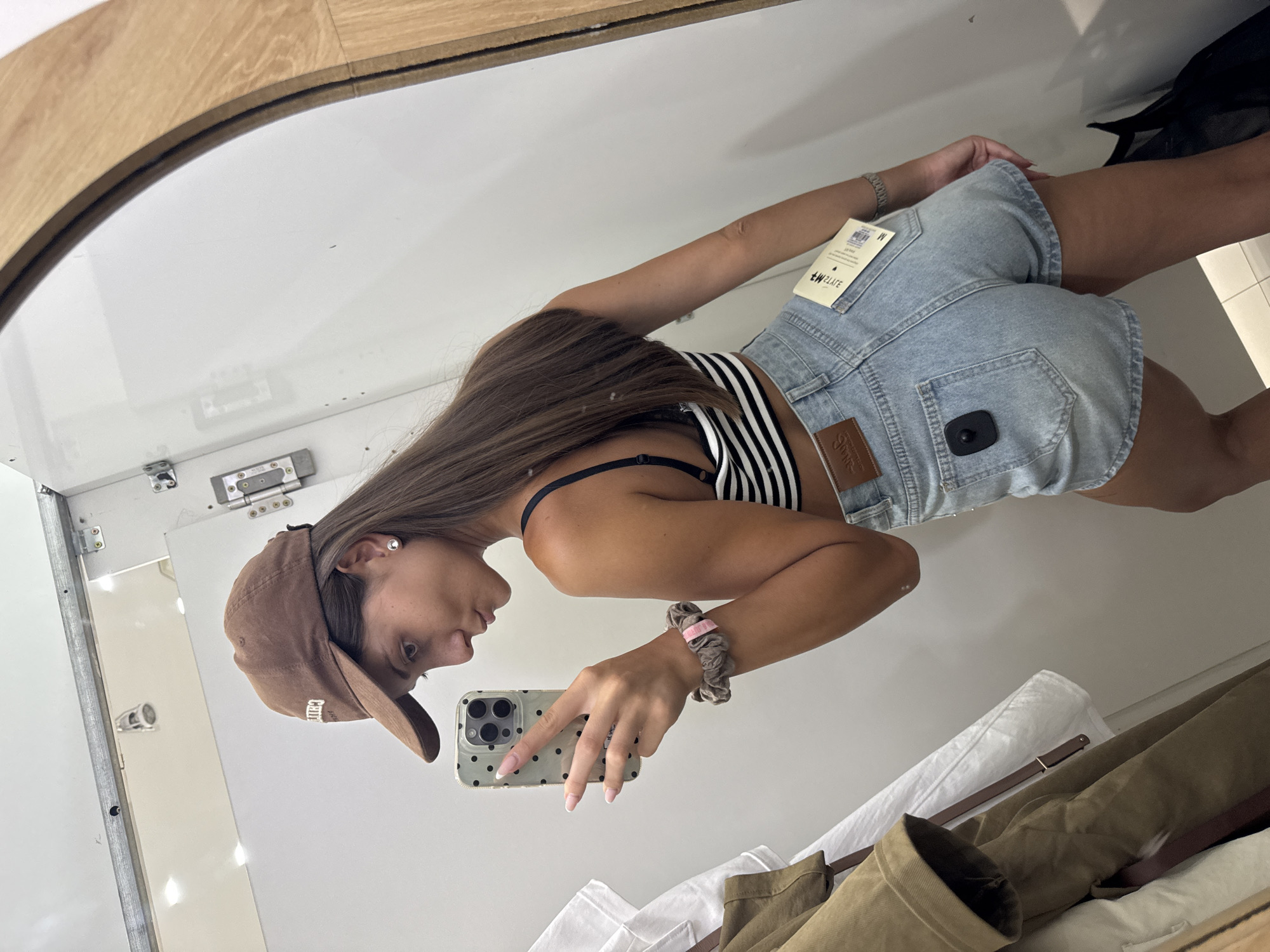 A young woman wearing a baseball cap, striped tank top, and denim shorts is taking a selfie in a dressing room. The dressing room has a mirror with a light wooden frame. Clothes hang on a rack below the mirror. The woman is holding a phone in her right hand, which has a scrunchie around the wrist. There is a tag on the shorts. The location is not discernible from the image.
