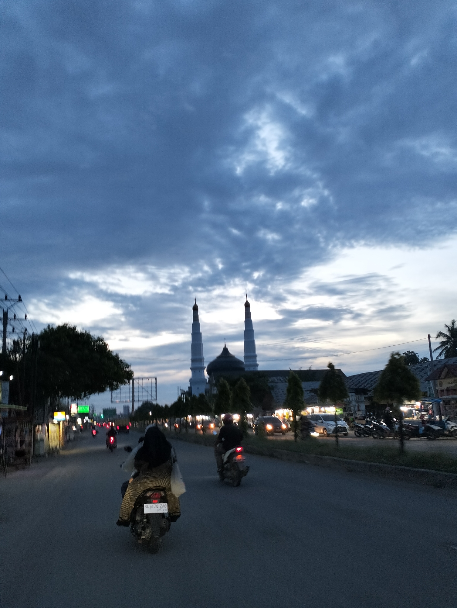 The photograph captures a twilight scene in Keudematangglumpangdua, Indonesia. Dominating the background is a mosque featuring a central dome and two tall minarets. The sky is filled with heavy, dark clouds, with some lighter patches hinting at the setting sun. A wide road extends into the distance, with streetlights illuminating the edges. Two motorcycles are visible on the road, one closer to the camera carrying two individuals, and the other further away with a single rider. Cars and additional parked motorcycles are present along the road. Trees and buildings line the sides of the road. Power lines are visible on the left side of the frame. The overall scene conveys an atmosphere of dusk settling over a town with moderate traffic.
