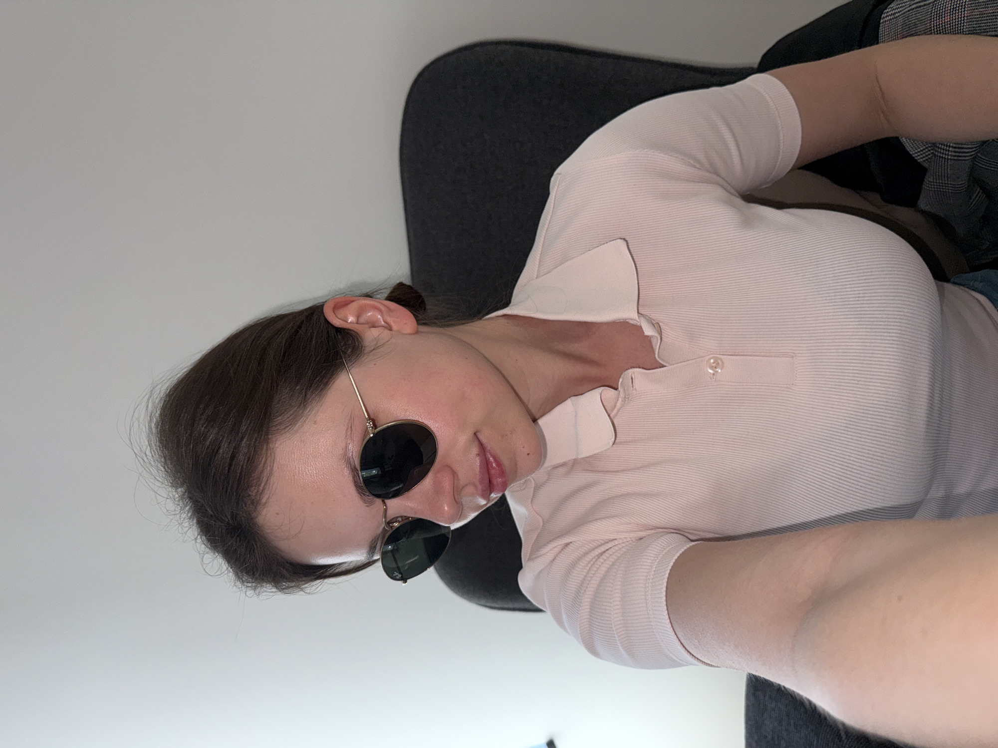 A young woman with dark hair pulled back is sitting in a dark gray chair indoors, wearing sunglasses and a light pink collared shirt. She is likely facing a white wall. The location is unknown and can't be determined to be Belgrade, Serbia.