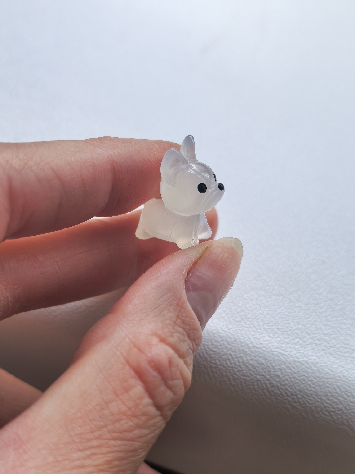 A person is holding a small, translucent figurine of a dog. The dog appears to be white or light gray, with black eyes and nose. The background is a plain white surface. The image does not provide information about the city of Sterlitamak, Russia.
