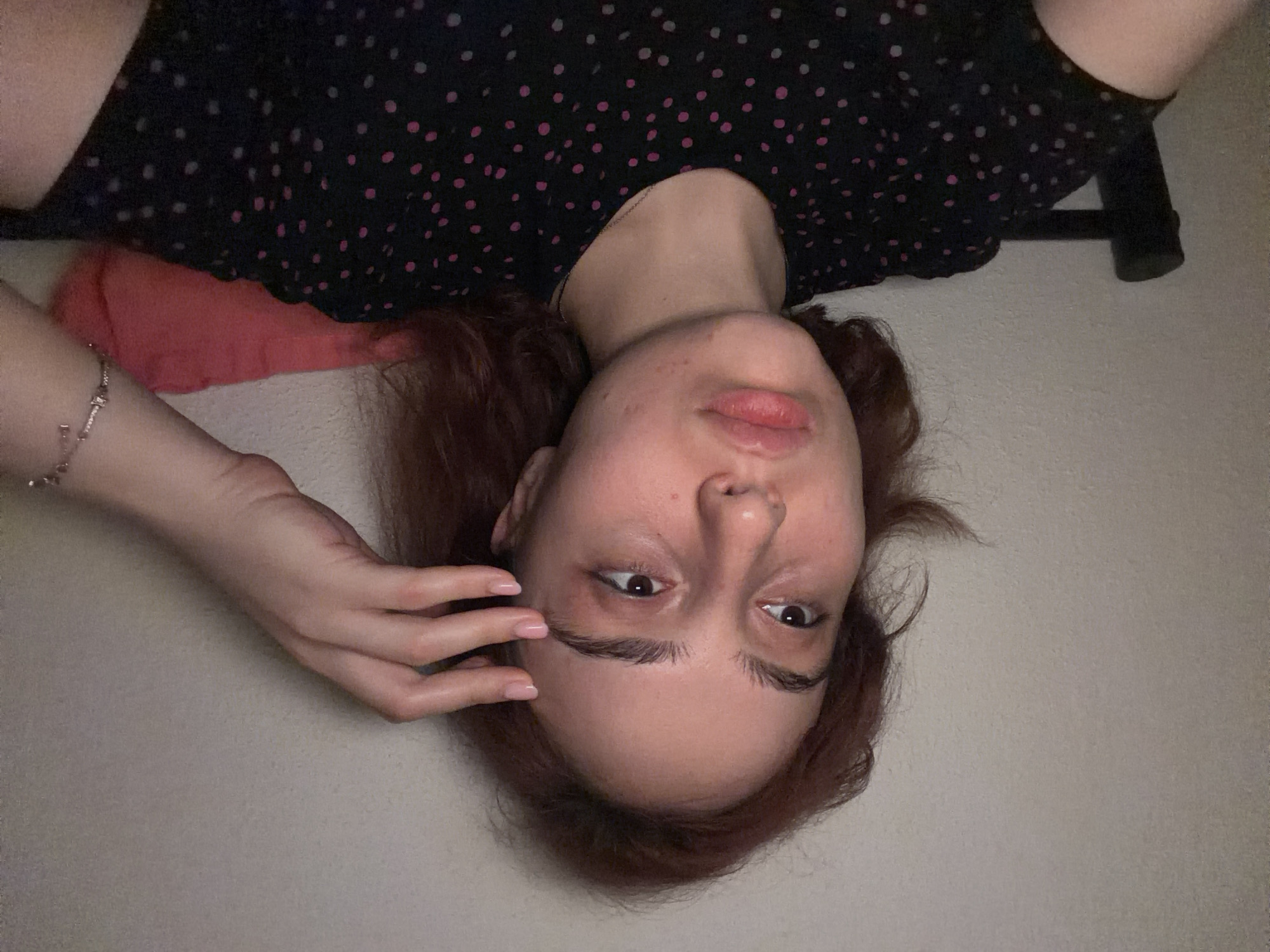 A young woman with reddish-brown hair is shown in a medium shot, looking off to the left. She has her right hand on her forehead, touching her temple. She is wearing a black t-shirt with pink polka dots. A pink pillow is visible behind her, and she is seated in a dark-colored chair. The background is a plain, textured wall. The lighting appears to be artificial and casts a warm tone on her face. Her expression is neutral, with her lips slightly parted. She is wearing a bracelet on her left wrist. The overall scene suggests an indoor setting, possibly a bedroom or living room. There is no indication of the image being taken in Strasbourg, France.