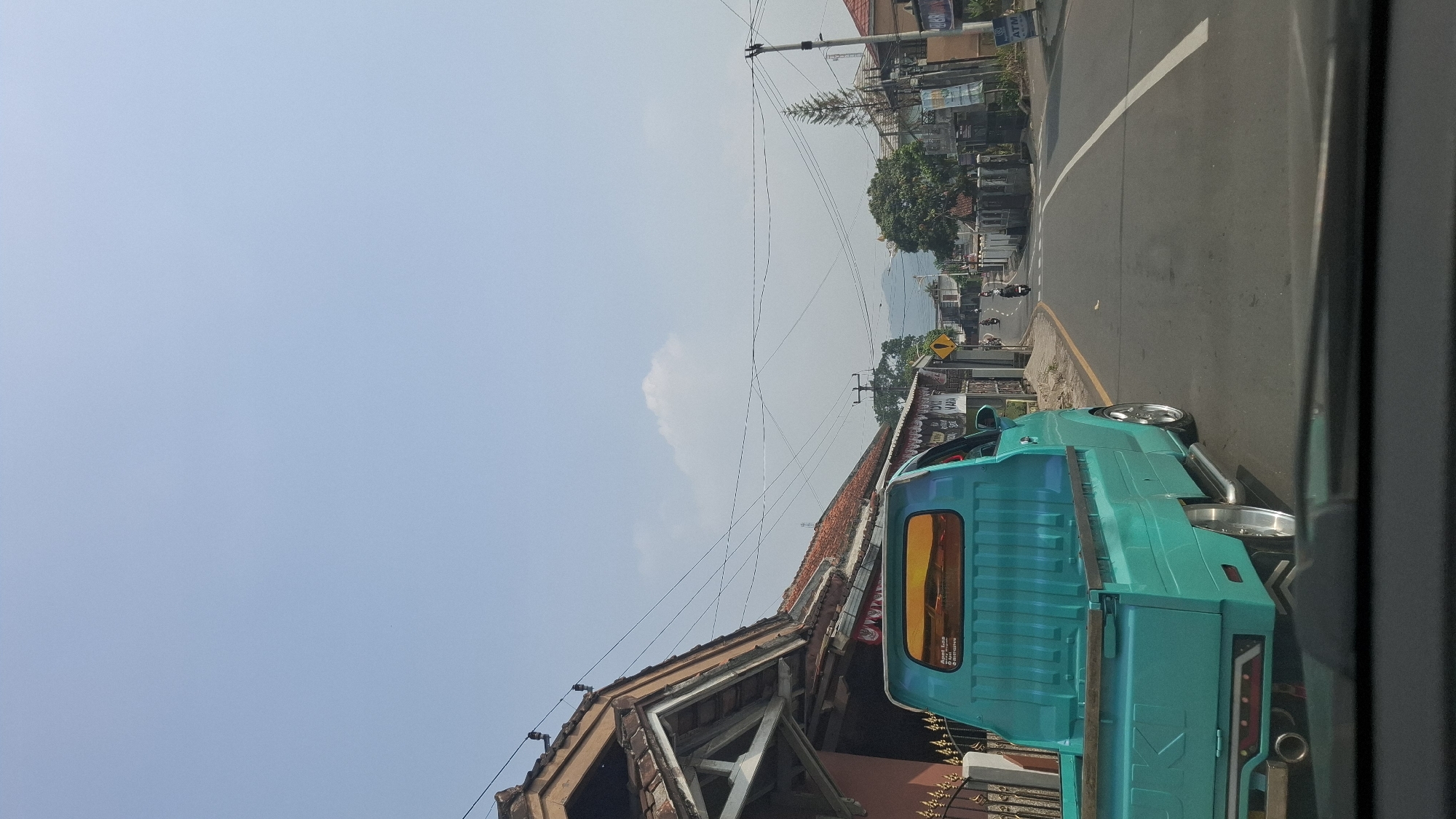A teal pickup truck is driving down a paved road in Cilaku, Indonesia. The truck is in the foreground, with its rear facing the viewer. The truck has custom rims and a lowered suspension. The road is lined with houses and shops. There are a few motorcycles and other vehicles on the road in the distance. The sky is clear and blue, with a few scattered clouds. The image appears to have been taken on a sunny day.