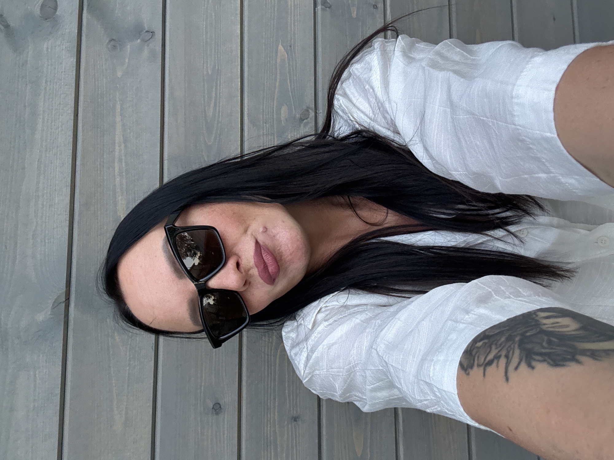 A woman with long, dark hair is wearing dark sunglasses and a white shirt in front of a grey wooden plank wall. The woman has a tattoo on her right arm. The scene's location in the city of Chaikovsky, Russia, cannot be determined from the image.