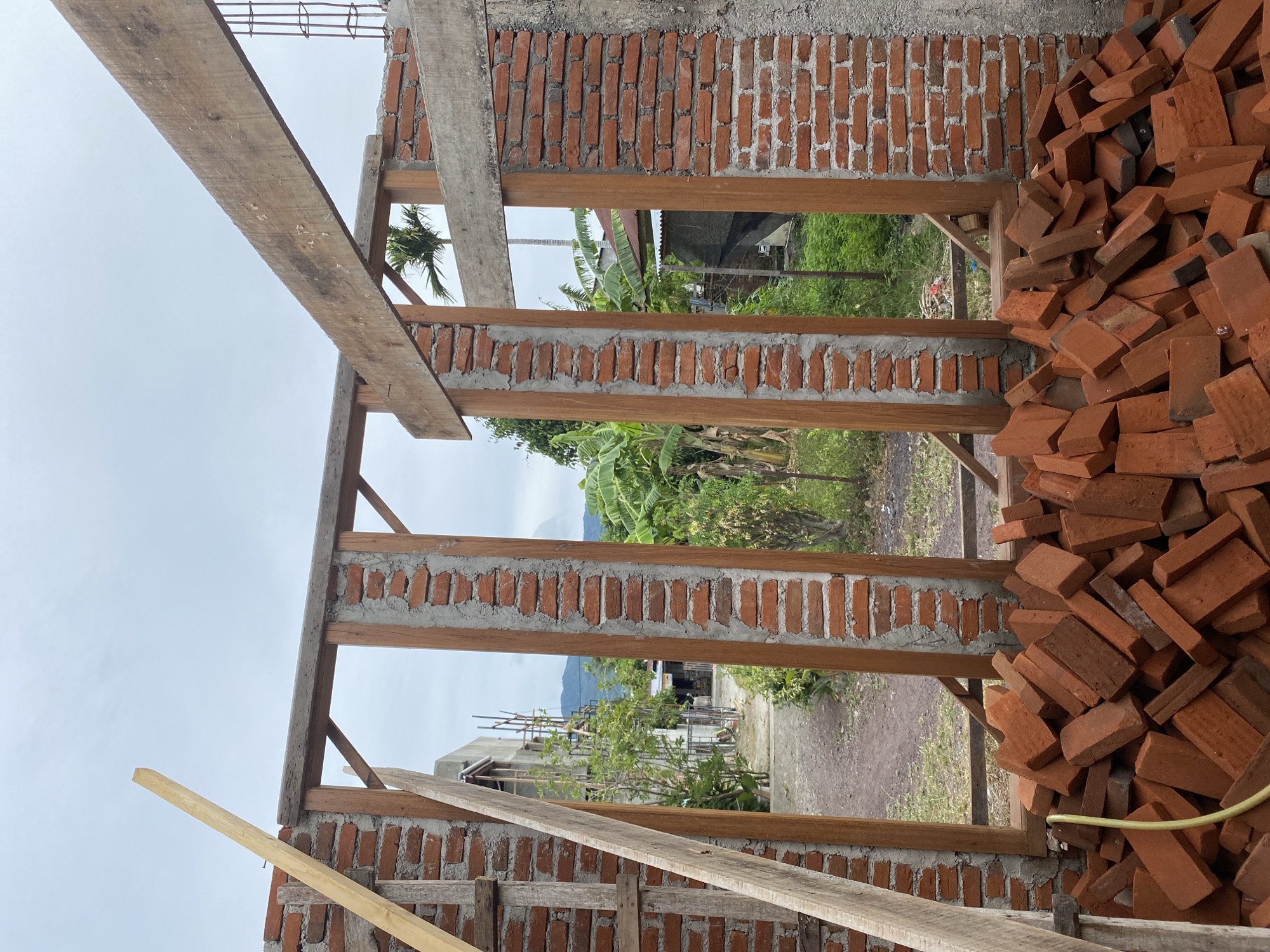 This image depicts the construction of a building in Banda Aceh, Indonesia. The foreground is dominated by a large pile of red bricks, with a yellow hose visible among them. The structure itself is partially built, featuring brick walls with wooden framing around window and door openings. Exposed concrete beams and rebar indicate ongoing construction. In the background, a dirt path leads towards other buildings and lush green vegetation, including banana trees and palm trees. The sky is overcast. The scene is a typical construction site in a tropical environment.