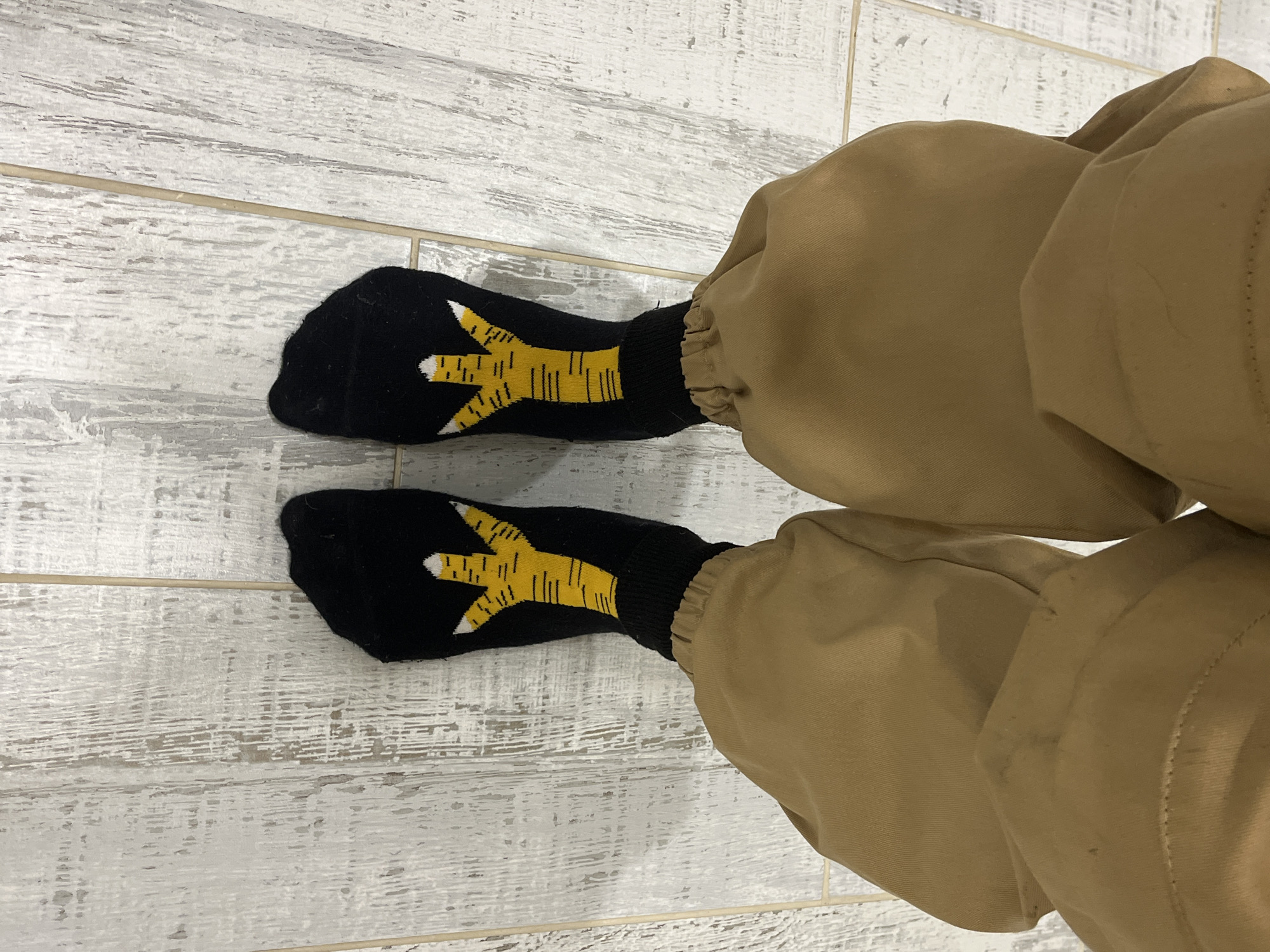 A first-person perspective shot shows two feet wearing black socks with a yellow chicken-foot pattern. The socks are partially covered by tan pants. The feet are positioned on a floor made of light-colored wooden planks. The overall lighting appears to be indoor ambient light.