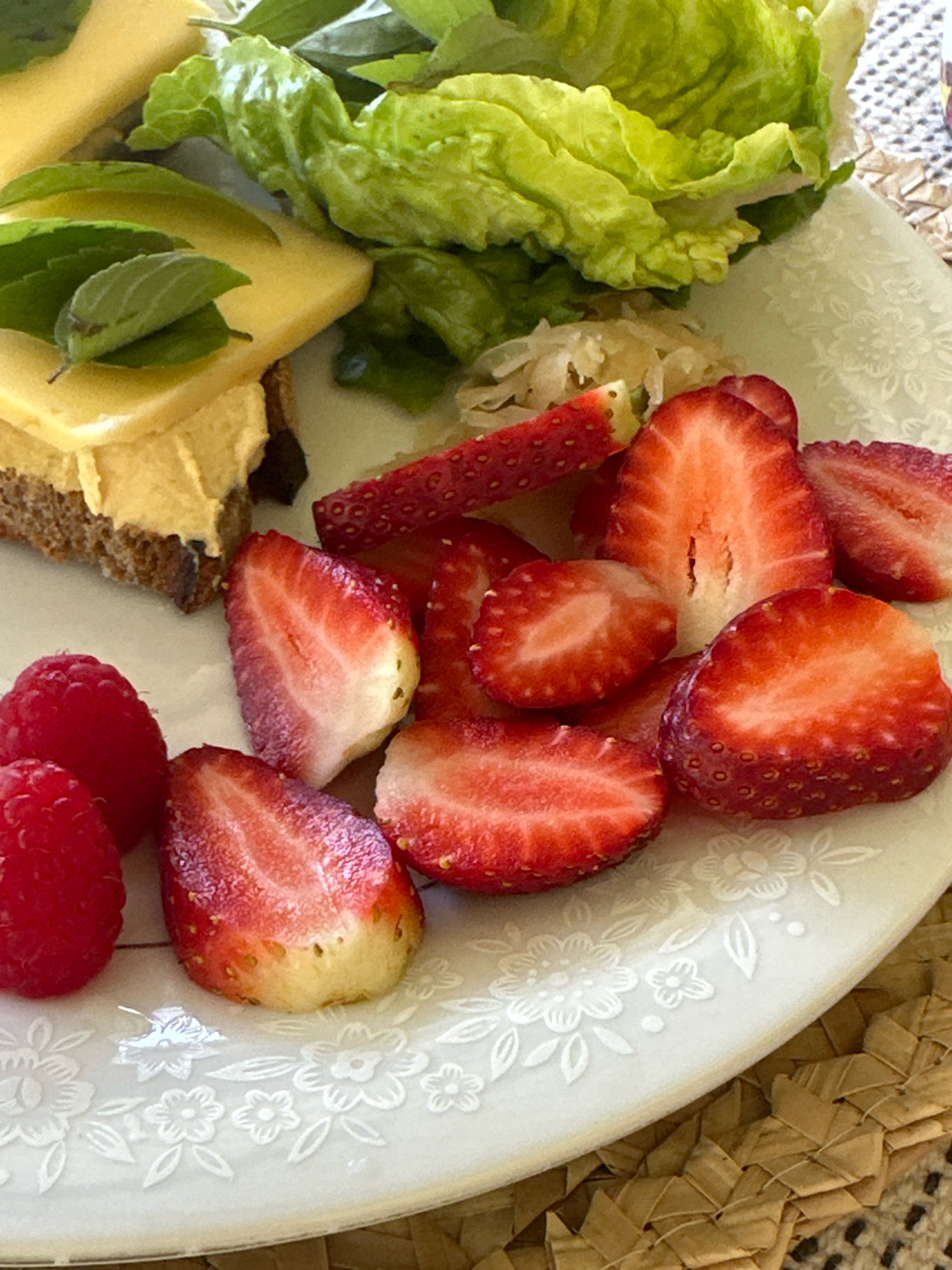 In Thora, Australia, a white ceramic plate with a floral embossed pattern holds a breakfast spread. Sliced strawberries and raspberries are arranged on the right side of the plate. To the left, a slice of bread is topped with what appears to be hummus or a similar spread, topped with a slice of cheese and garnished with fresh mint leaves. A small mound of what looks like sauerkraut is visible between the cheese and the strawberries. The plate rests on a woven placemat.