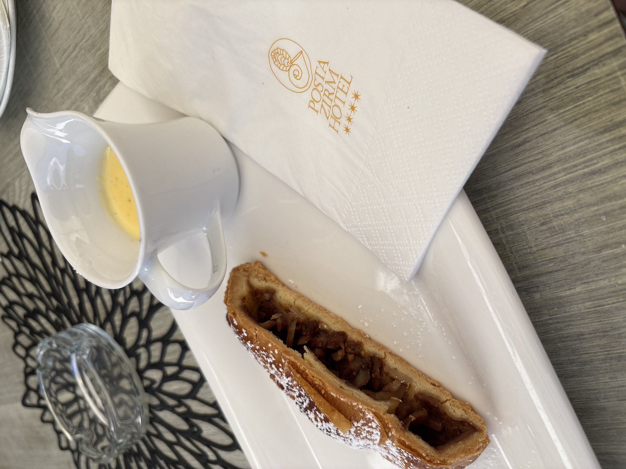 An overhead, slightly angled shot shows a white rectangular plate on a wooden table. On the plate rests a piece of apple strudel dusted with powdered sugar, a white creamer filled with yellow custard, and a white napkin with the gold "POSTA ZIRM HOTEL" logo and four stars. To the left of the plate, on a black woven placemat, sits a glass ashtray. The overall scene depicts a dessert setting.