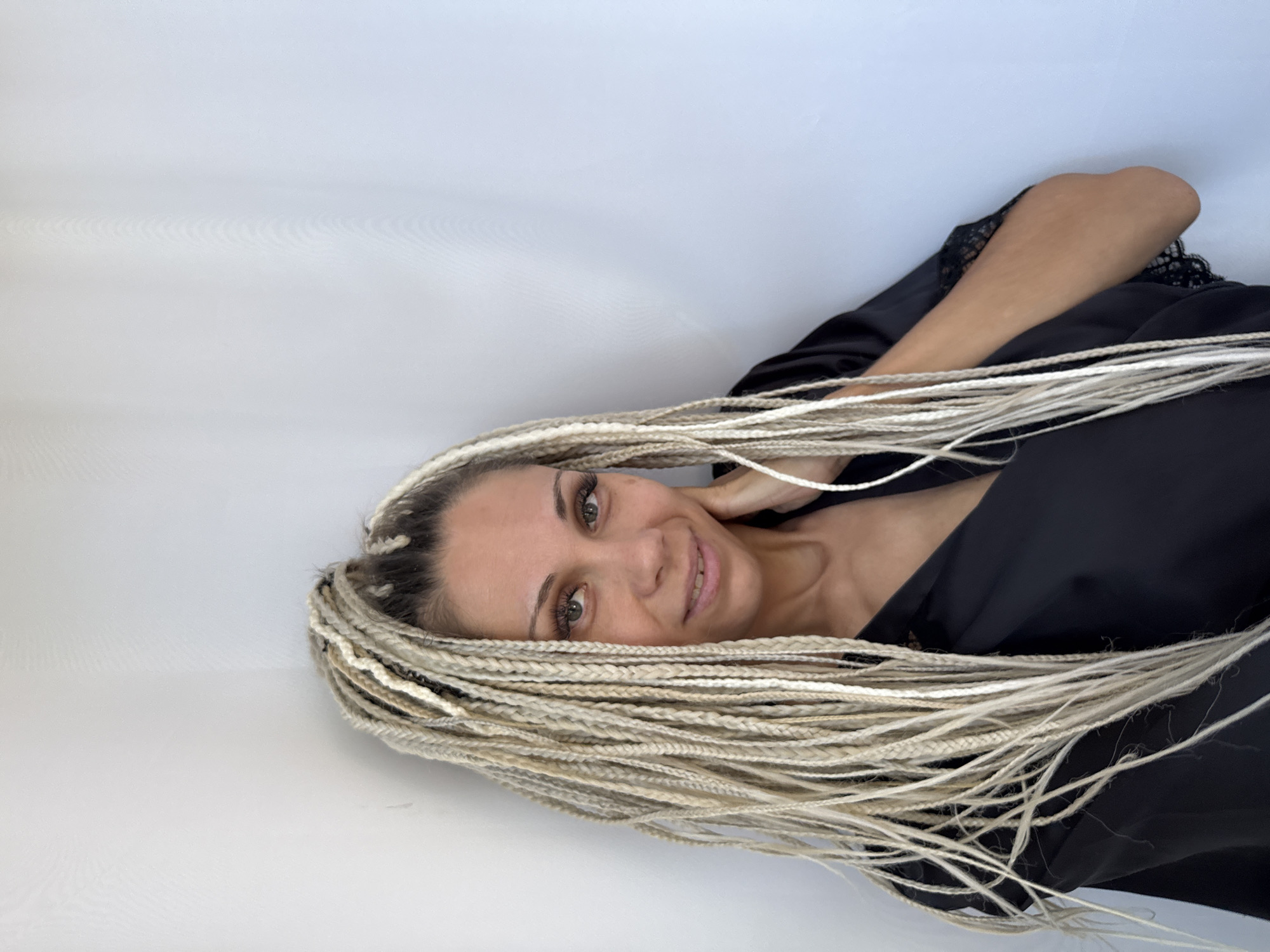 A woman with blonde braids is posing indoors against a white wall. She is wearing a black robe and has her hand on her neck. The lighting suggests it is daytime, possibly from a window or studio lighting. The image is a close-up shot focusing on the woman's upper body and face. There is no information in the image to suggest this is in the city of Rusanovo, Russia.