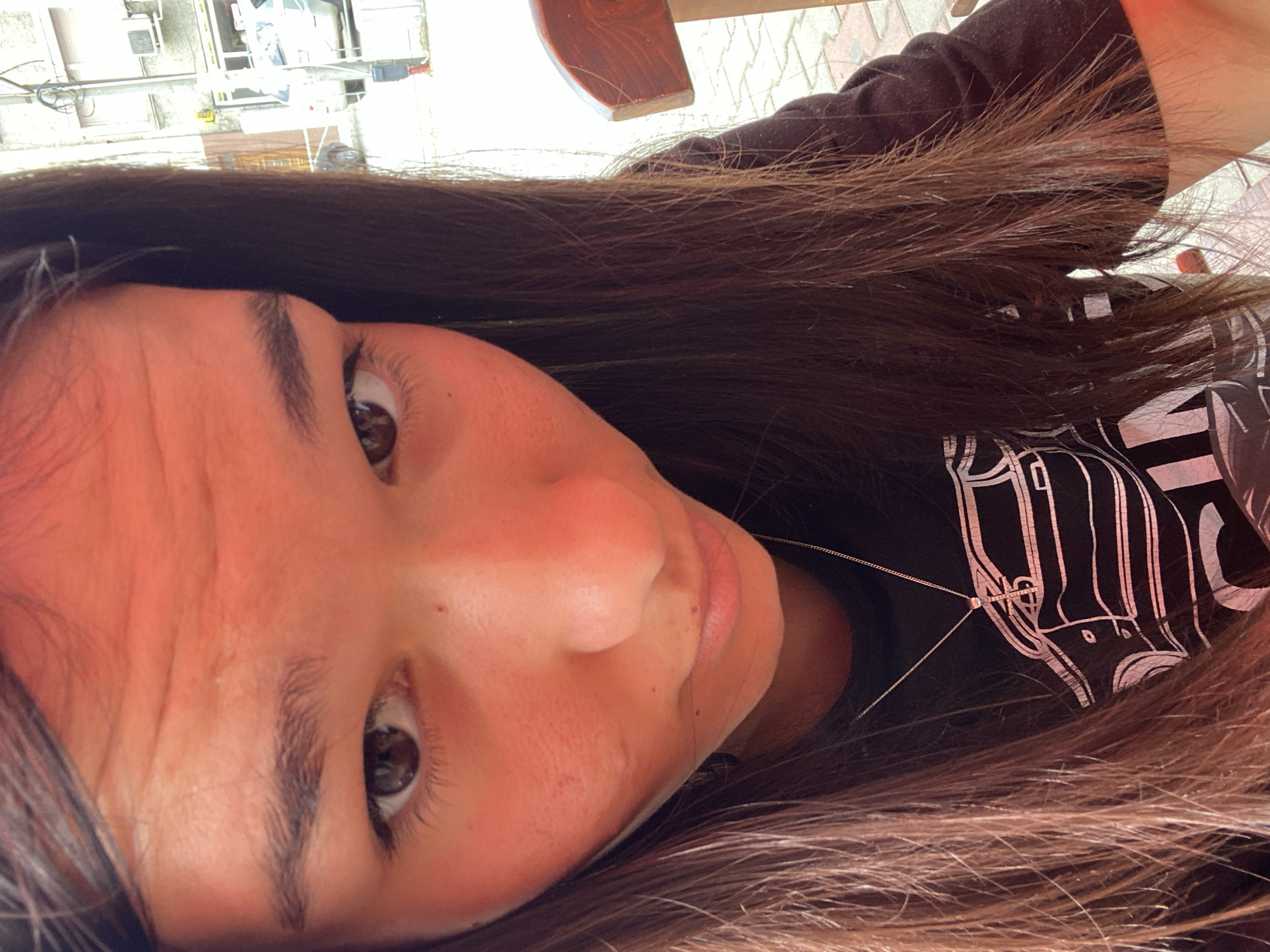 A young woman with long, dark brown hair is captured in a close-up, angled selfie. Her face is illuminated with a warm, orange light, likely from an awning or overhead structure. She is wearing a dark t-shirt with a white graphic of a car on it and a delicate silver necklace. The background shows a blurred urban scene with buildings and a vehicle, suggesting an outdoor setting. The image appears to have been taken during the daytime. The location is identified as Sevlievo, Bulgaria.