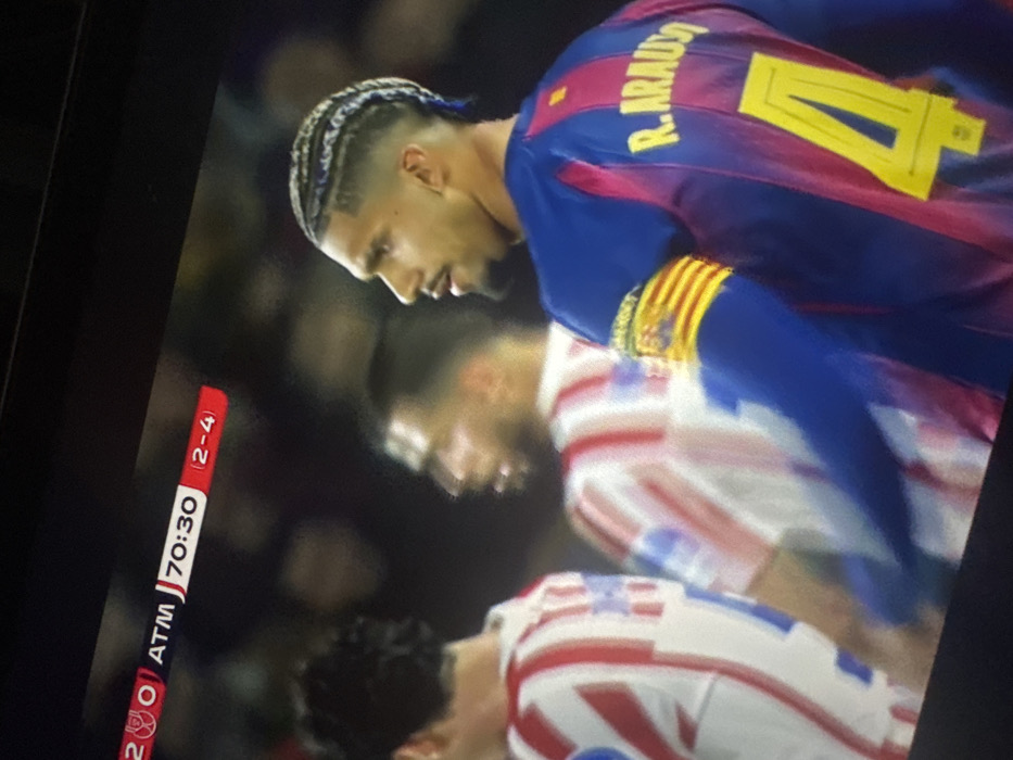 The image displays a photograph of a television screen showing a professional soccer match, observed in Gainesville, United States of America. In the foreground, three male soccer players are visible on a field. The most prominent player, positioned on the right, wears a blue and red striped jersey with "R. ARAUJO" and the number "4" in yellow on the back. He has braided hair with lighter strands, a fade haircut, a beard, and a mustache. A captain's armband, yellow with red stripes, is visible on his left bicep. He is looking towards the left side of the frame. Two other players are partially visible behind him, both wearing red and white striped jerseys; one is in the middle ground, slightly out of focus, and the other is largely obscured on the left.
A digital score bug is present in the upper left portion of the screen. It displays "2 0" next to a logo resembling Arsenal FC's cannon, "ATM J 70:30" indicating Atlético Madrid and the match time of 70 minutes and 30 seconds, and a red box on the right showing "(2-4)". The background is blurred, suggesting a stadium environment with spectators. No specific interaction between the players is depicted; they appear to be standing on the field during play.