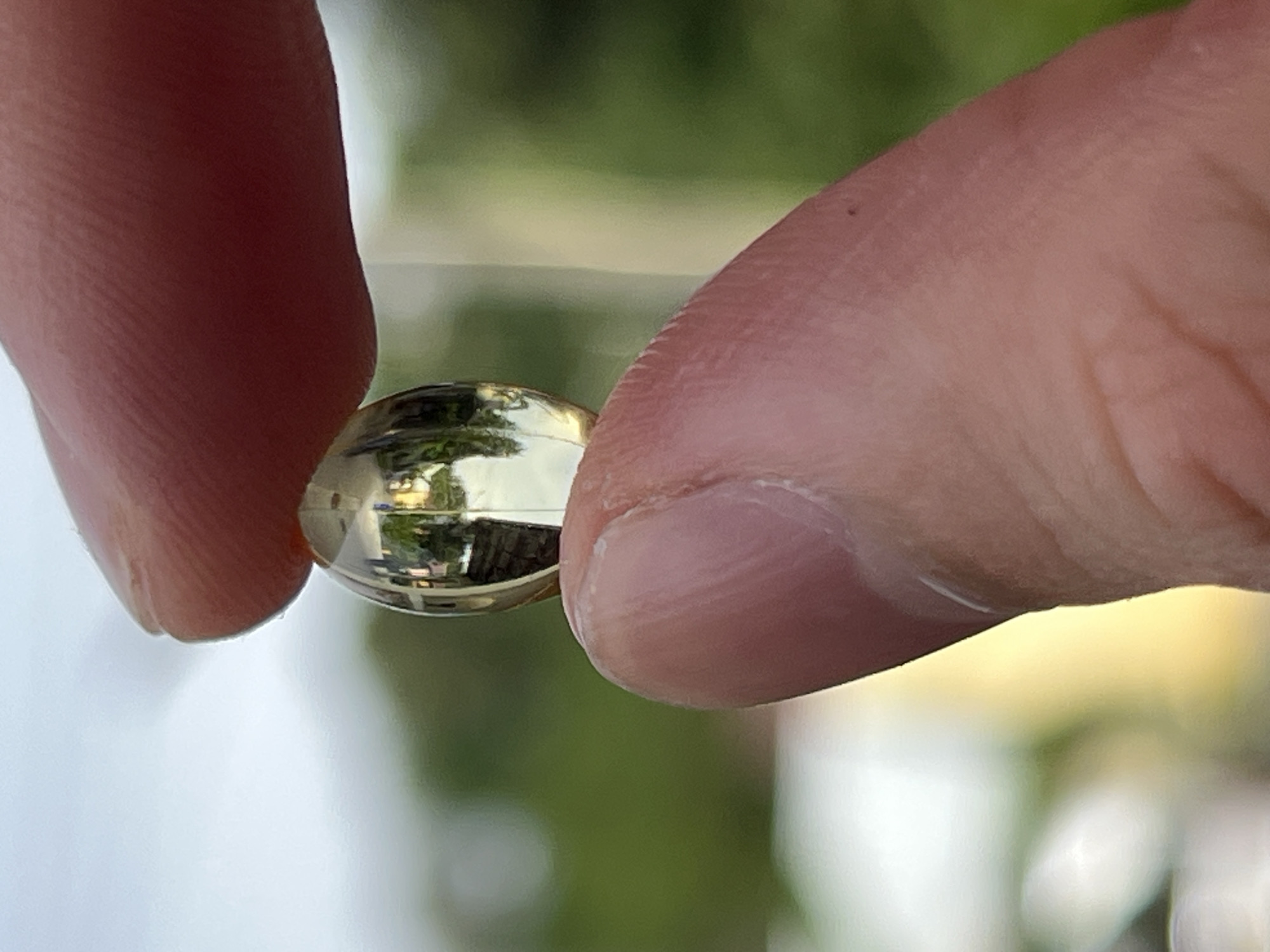 A close-up shot shows a person's thumb and index finger holding a clear, oval-shaped capsule. The capsule is transparent and appears to contain a yellow liquid or gel. The background is blurred, showing green foliage and a light-colored sky, suggesting an outdoor setting. Reflections within the capsule hint at surrounding objects and light sources. The image is taken from a slightly low angle. The setting is outside in a residential area in Choeng Thale, Thailand.
