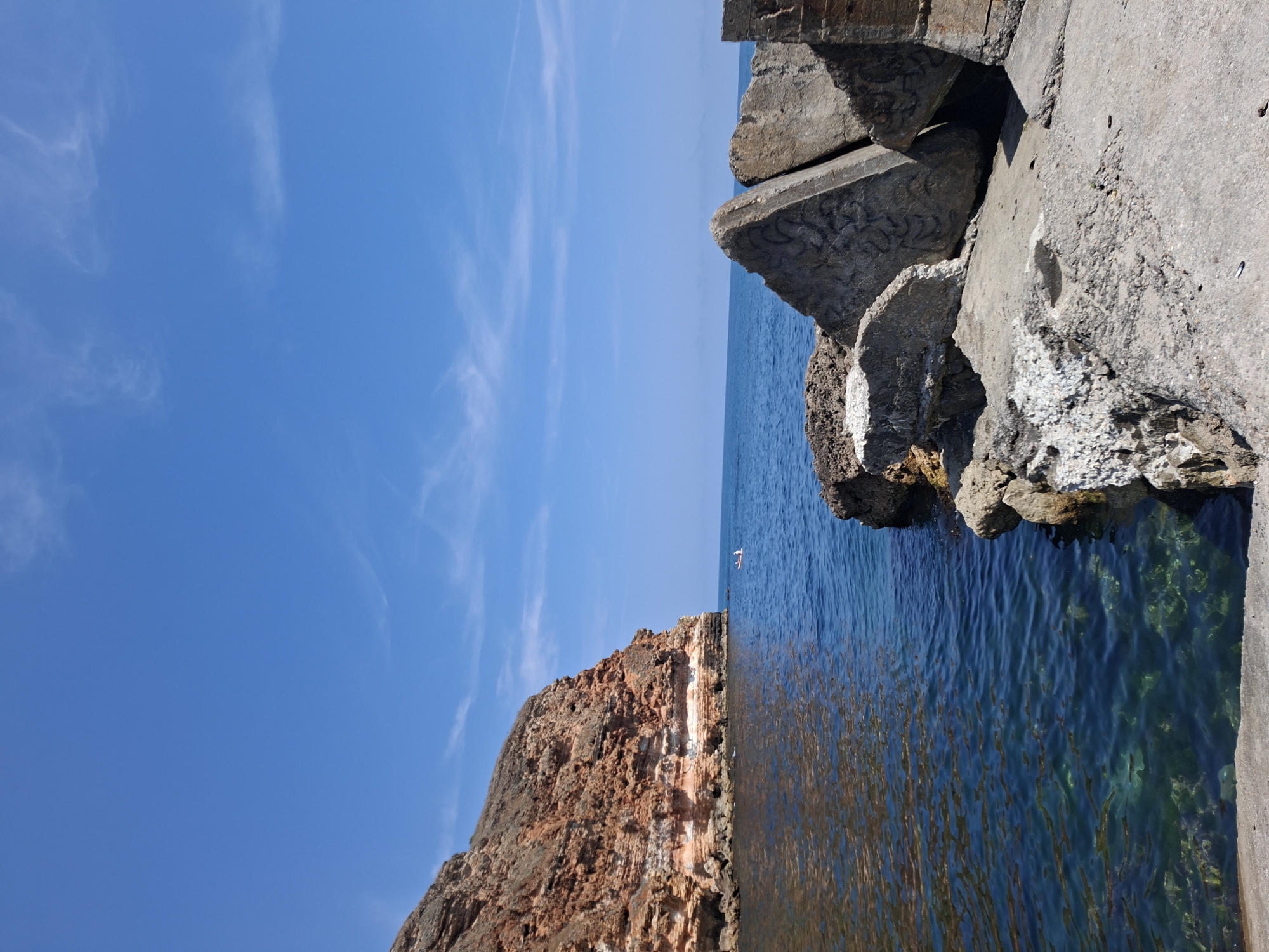 The image captures a scenic view of the Black Sea coast near Balgarevo, Bulgaria. The foreground is dominated by a concrete structure, likely part of a breakwater or pier, with large, angular concrete blocks piled on top of one another. Some of these blocks are weathered and appear to have graffiti. To the left, the clear, blue water of the sea laps against a rocky shore and a towering, reddish-brown cliff face. The cliff exhibits distinct geological layers and shows signs of erosion. The water itself is remarkably clear, allowing glimpses of the seabed beneath. In the distance, a small white boat with a person in it is visible on the calm sea. The sky above is a vibrant, cloudless blue, with a few wispy cirrus clouds streaking across it. The overall impression is one of a rugged and beautiful coastal landscape on a sunny day.