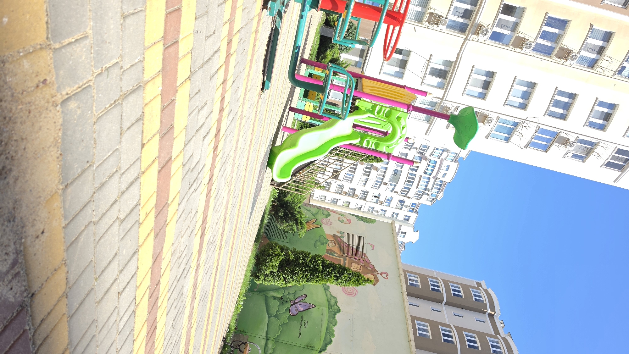 An outdoor shot captures a vibrant playground with a bright green slide and red carousel, set against a backdrop of modern apartment buildings and a clear blue sky. The playground ground is paved with a patterned mix of gray, yellow, and red interlocking stones, with a few small green bushes and trees surrounding it. A colorful mural adorns one of the building walls, depicting a whimsical house with a heart-shaped chimney and butterflies. The buildings are tall and contemporary, with numerous windows and air conditioning units visible. The lighting suggests a sunny day.