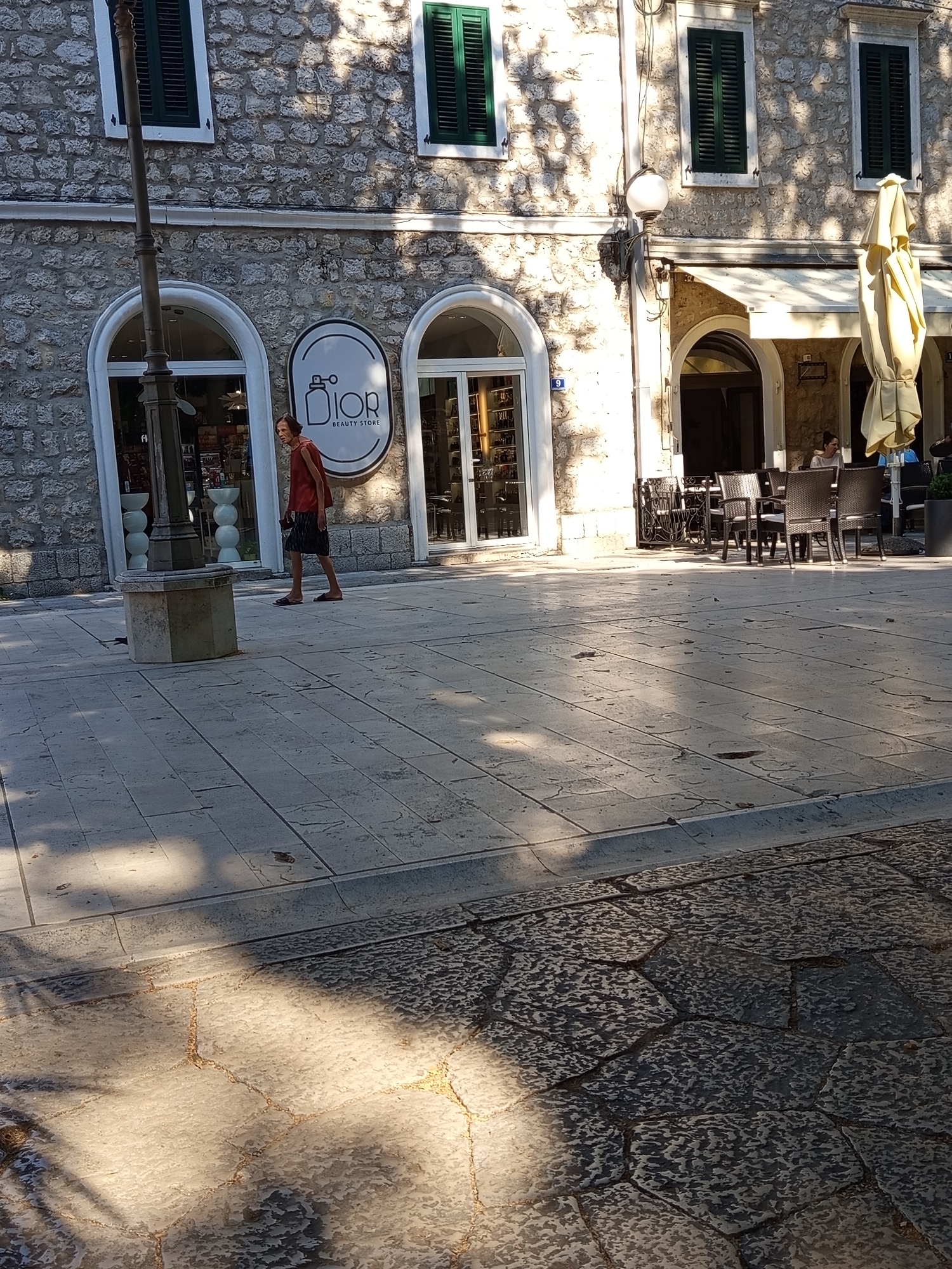 This image depicts a street scene in Trebinje, Bosnia and Herzegovina, likely near the Old Town area given the traditional stone architecture. A person wearing a sleeveless reddish-orange top and a dark skirt is walking on a paved sidewalk. To their left, a stone pedestal with a dark metal fixture stands on the sidewalk. Behind the person, a building features an arched entrance with the Dior Beauty Store logo displayed above it. Further to the right, an outdoor seating area for a cafe is visible with tables and chairs under a yellow umbrella. Sunlight casts shadows across the paved ground.