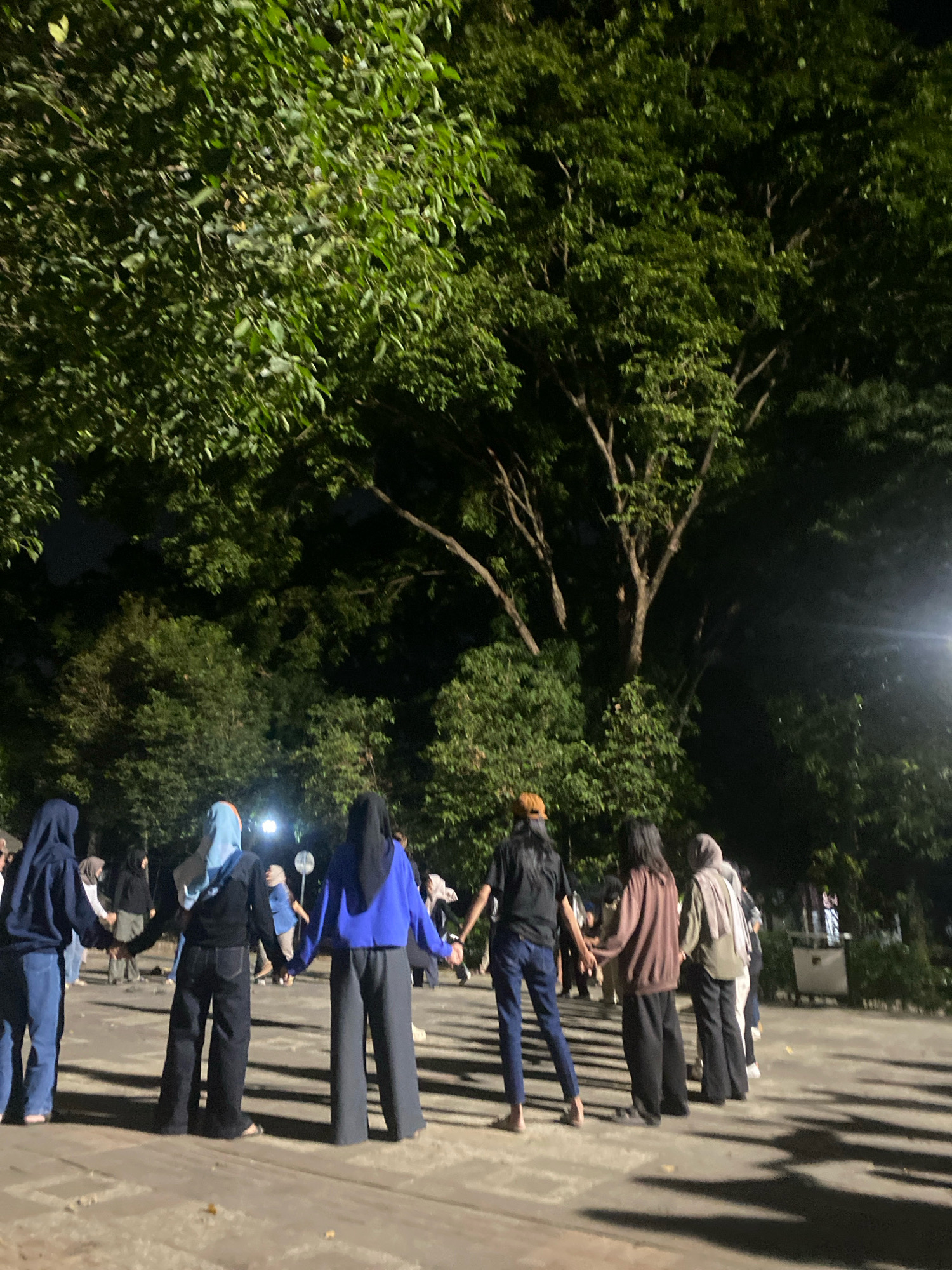 A group of at least ten individuals, predominantly women, are holding hands and forming a circle outdoors at night. They appear to be engaged in a communal activity or game. The setting is an open paved area, possibly a park or public square, with mature trees providing a dense canopy overhead. The lighting is dim, with some artificial light sources visible, casting shadows on the ground. The presence of trees and the general ambiance suggest a location in a city environment with green spaces, which could be found in various parts of Semarang, Indonesia, such as Kota Lama or an outdoor recreational area. The individuals are dressed in casual attire, with several wearing headscarves.