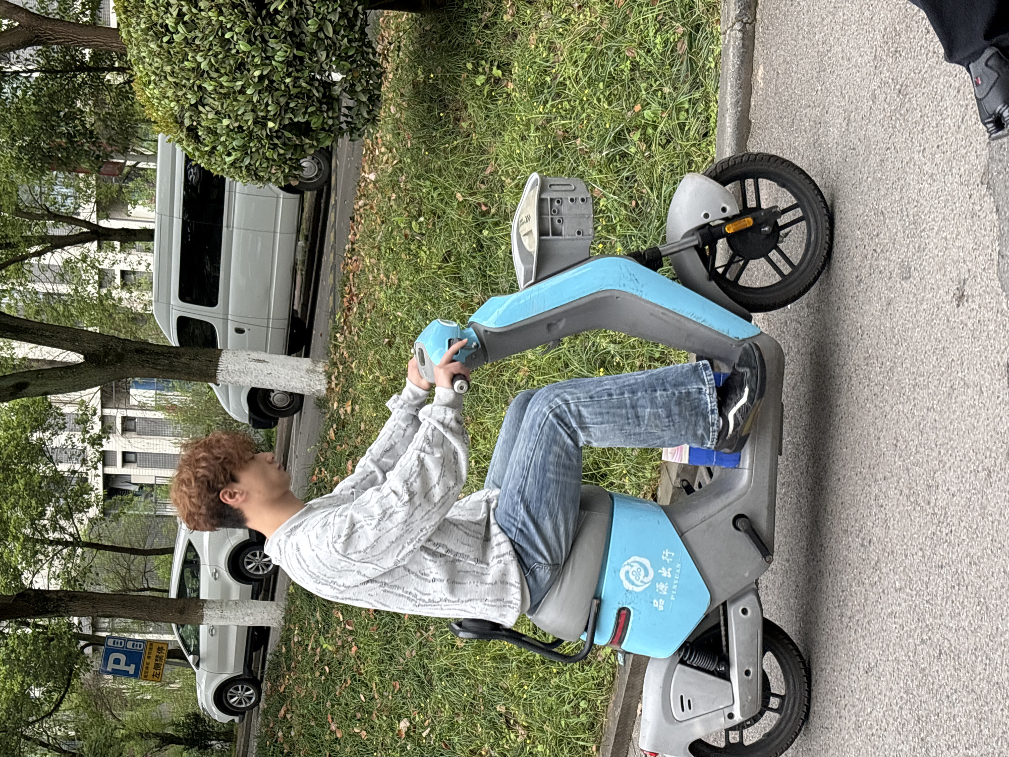 A male individual with light-brown, curly hair is depicted riding a light blue and grey electric scooter on an asphalt road in Jiangjin, China. The individual wears a light grey long-sleeved shirt with a textured pattern, blue jeans, and black sneakers with white accents. His hands are on the handlebars, and his feet are positioned on the scooter's footrests.
The electric scooter features a blue main body, grey trim, a grey seat, and a front basket. The brand "品源出行" (Pinyuan Chuxing) is visible on its side. To the right, a small portion of a black shoe belonging to an unseen individual is visible on the road surface.
The background consists of a grassy area to the immediate left and right of the road, featuring green grass, scattered dried leaves, and small yellow flowers. Several trees with white-painted bases and green foliage are present. Behind the trees, a white SUV is parked on an elevated section, and further back, a light grey minivan is also parked. A blue rectangular parking sign with Chinese characters "此处停车 左侧禁停" (Parking here, No parking on the left side) is visible on the left. Light-colored multi-story buildings are partially visible through the trees in the far background. The sky is overcast, suggesting diffused lighting.