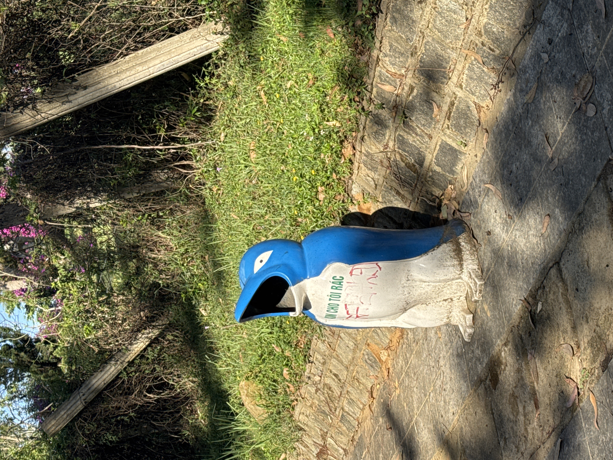 A horizontal eye-level outdoor shot depicts a blue-and-white penguin-shaped trash receptacle situated on a stone-paved surface within a park, presumed to be in Lam Vien – Da Lat, Vietnam. The receptacle has the phrase "KIN CHO TOI RAC" written on its side.
The paved surface consists of interlocked stone squares, marked with scattered brown leaves and minor vegetation. Adjacent to the stone surface, there is a patch of green grass. Behind the grass, a fence-like structure composed of two parallel concrete beams is visible, partially obscured by dense foliage, branches, and a cluster of pink flowers.