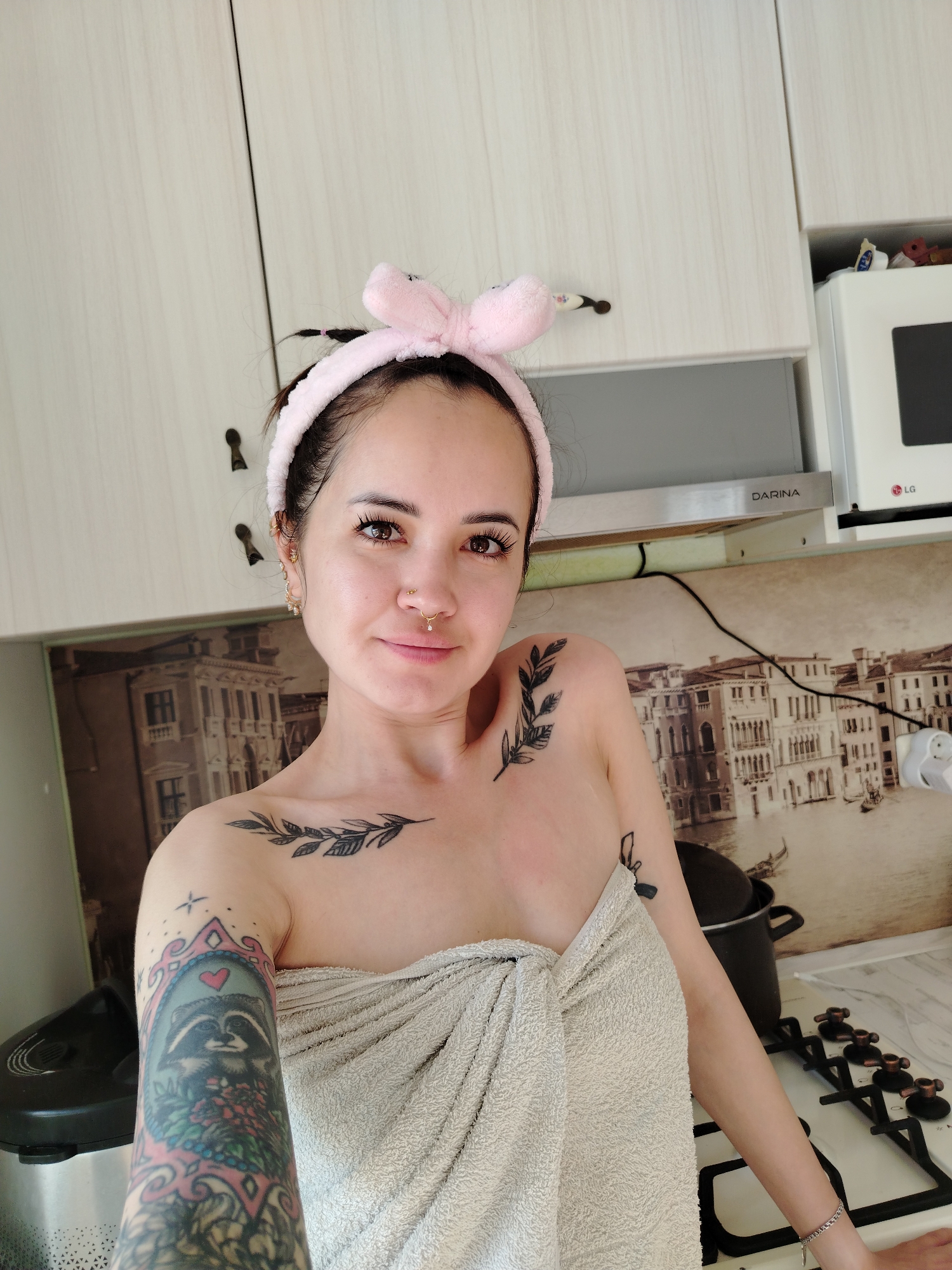 A young woman with dark hair, wearing a pink fluffy bunny ear headband and a towel wrapped around her chest, poses for a selfie in a kitchen. She has a nose ring and several tattoos on her chest, shoulder, and arm, including a raccoon in a frame. The kitchen features light-colored cabinets, a stovetop, and a wallpapered backsplash depicting buildings and water, resembling Venice. An LG microwave is visible on the top right.