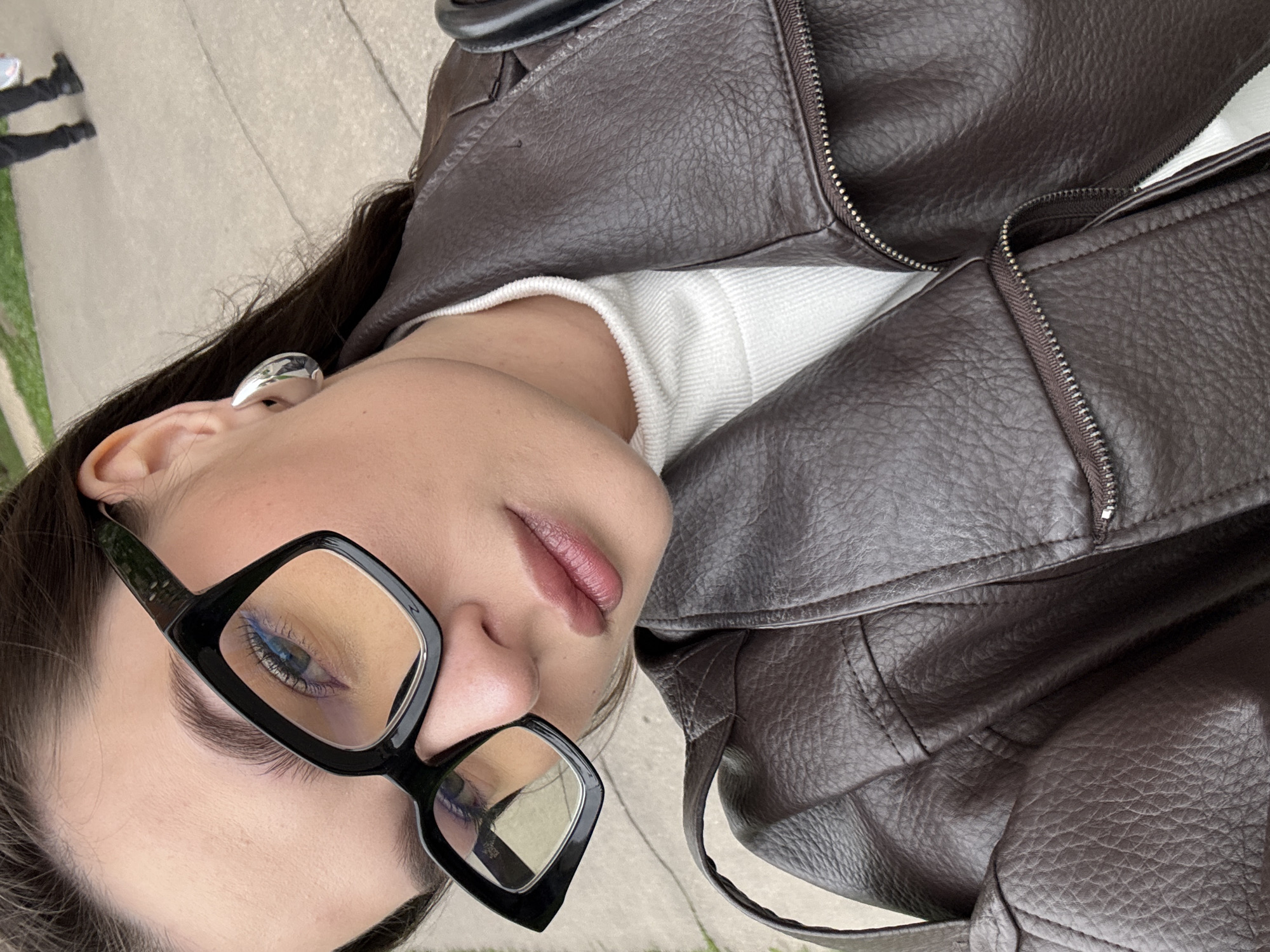 A close-up, angled shot captures a woman wearing black square-framed glasses, a white sweater, and a brown leather jacket. The focus is on her face and upper torso. Part of a sidewalk and some greenery are visible in the background, suggesting an outdoor urban setting. There is no information to indicate the location is Vnukovo, Russia.