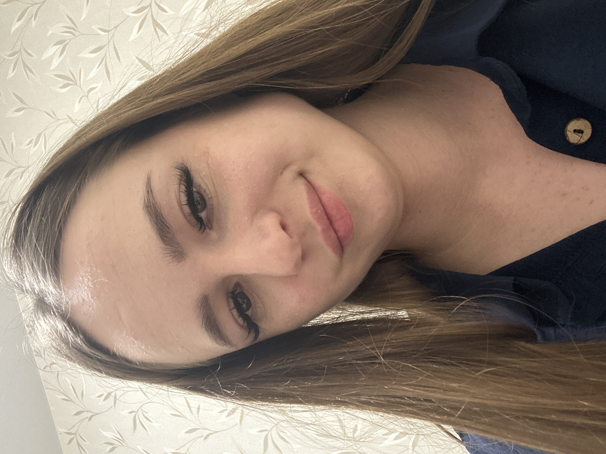 A portrait of a woman with long brown hair, wearing a dark blue collared shirt with a tan button, posing in front of a patterned off-white wall. The location is Unknown, Unknown.