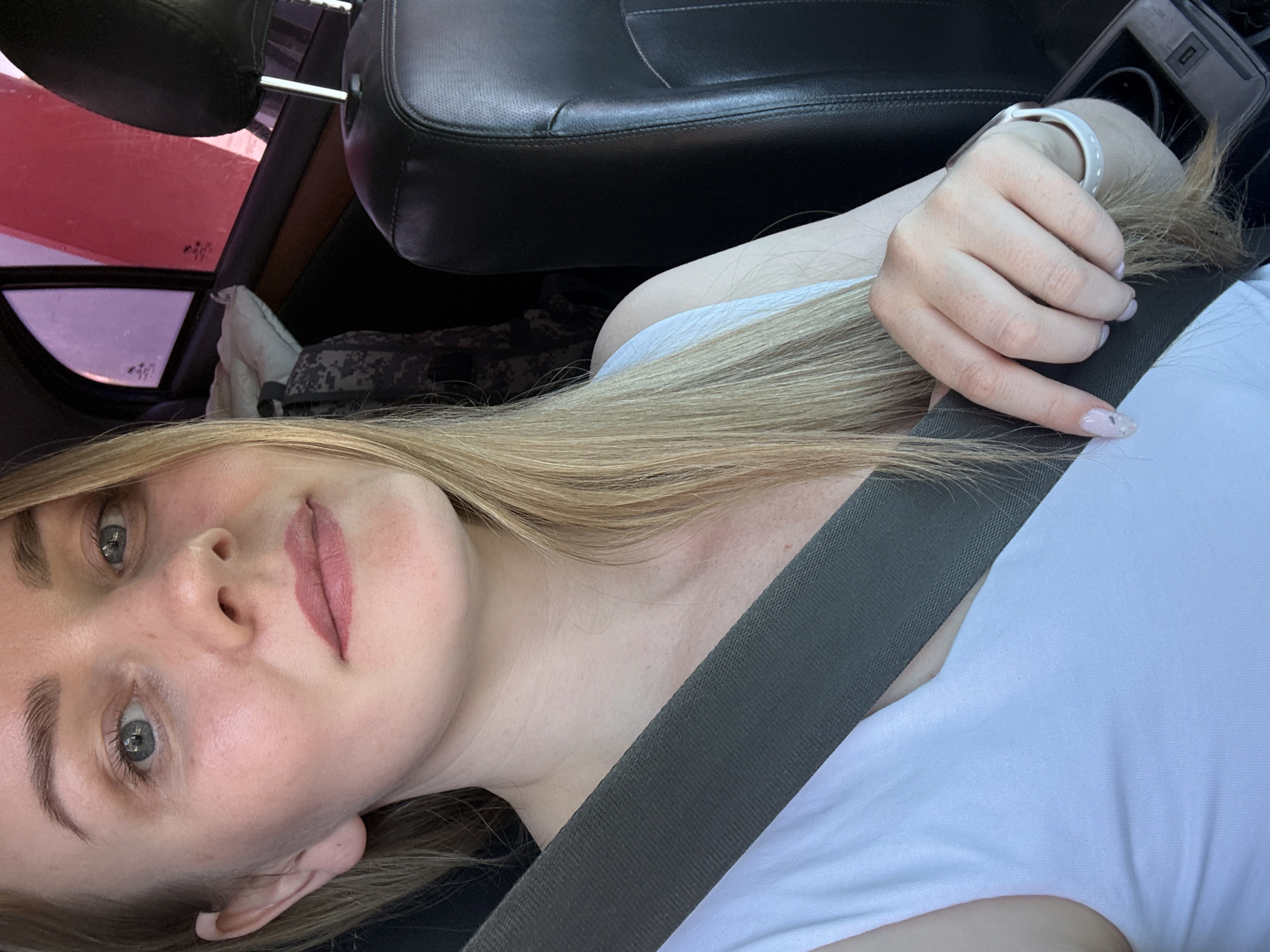 A blonde woman is pictured inside a car, wearing a white shirt and a seatbelt. Her left hand rests on the seatbelt, and she is wearing a white bracelet. The car has black leather seats and the background shows parts of the car's interior, including the dashboard and tinted windows. The location of the image is unknown, the city of Chaikovsky, Russia cannot be confirmed as there is no clear information about the location.