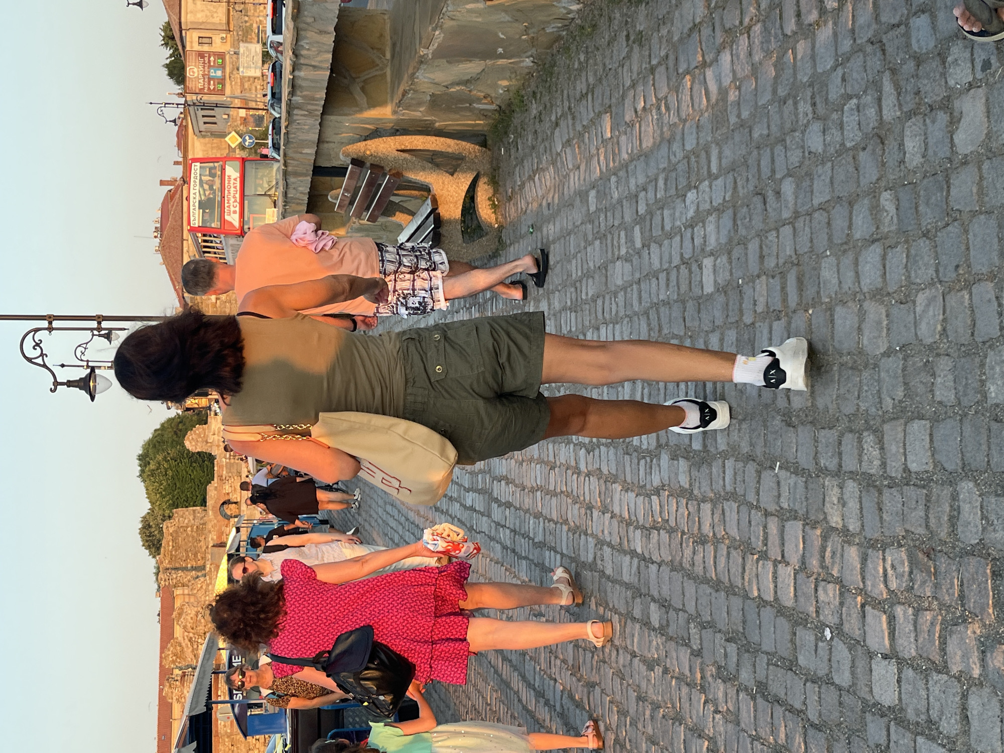 A group of people are walking on a cobblestone path in the city of Nesebar, Bulgaria. In the foreground, a woman wearing khaki shorts and a green tank top walks away from the camera, carrying a beige tote bag. To her left, a woman in a bright pink dress walks with a child. Further ahead, a man in a light pink t-shirt and patterned shorts walks with another person. In the background, a red double-decker bus with Cyrillic text is visible, along with a stone wall and ancient ruins. The sun is setting, casting long shadows on the path.