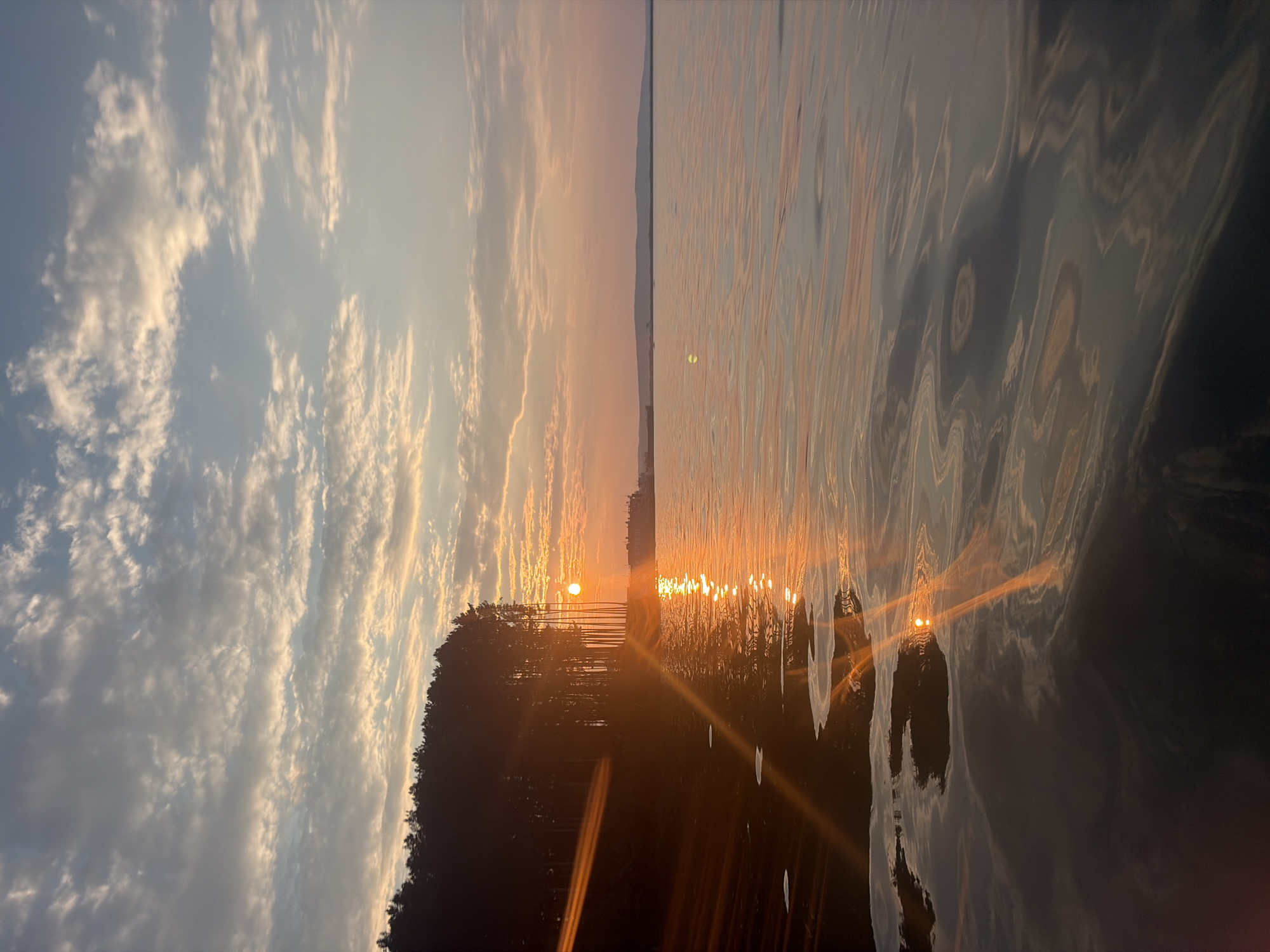The image depicts a sunset over a body of water, likely a lake or river, with a forested shoreline on the left. The sky is filled with wispy clouds, tinged with the warm oranges and yellows of the setting sun. The sun itself is visible, a bright orb reflecting on the rippling surface of the water, creating a path of light that stretches towards the viewer. The water is dark and reflects the colors of the sky and the silhouette of the trees. The overall scene evokes a sense of tranquility and natural beauty. The time of day is clearly sunset, given the colors of the sky and the sun's position. The weather appears clear with some cloud cover. The location, Chaikovsky, Russia, suggests a temperate climate and likely a natural, outdoor setting. There is no visible text in the image.