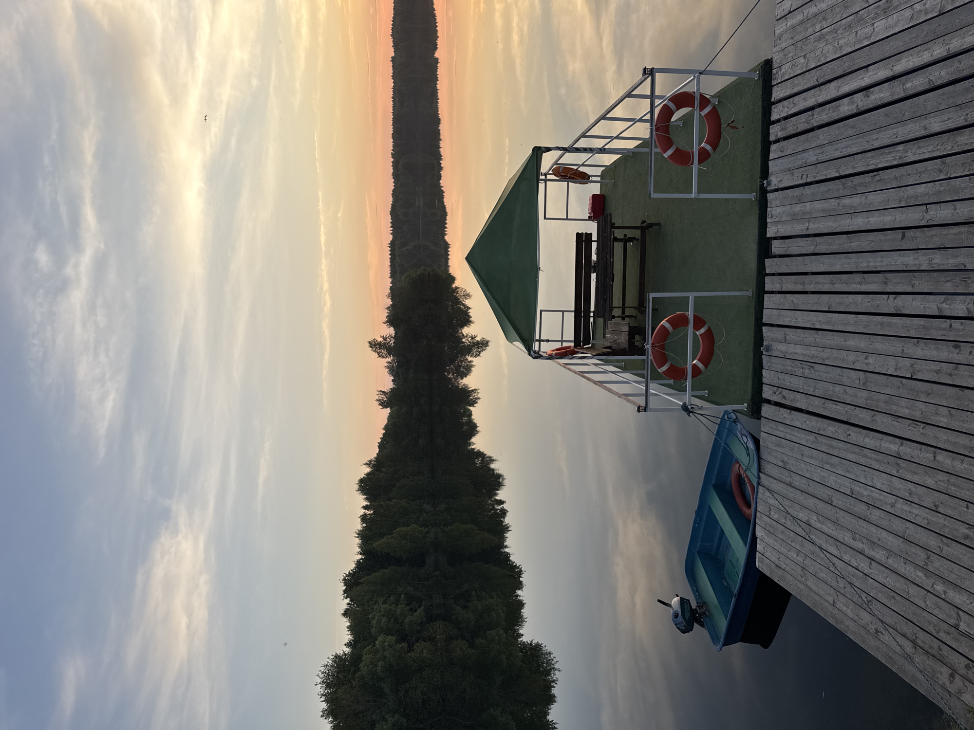 The image depicts a serene lake scene at dusk, likely near Kamysh, Russia. The sky is painted with soft hues of orange, yellow, and blue, with wispy clouds reflecting on the still water. A wooden pier extends into the lake, with a small blue motorboat tied to it. Further out, a green-roofed floating platform features a picnic table with benches and several life preservers. The opposite shore is lined with a dense forest, its dark silhouette contrasting with the colorful sky and its reflection mirrored in the water. There are no people visible in the frame.