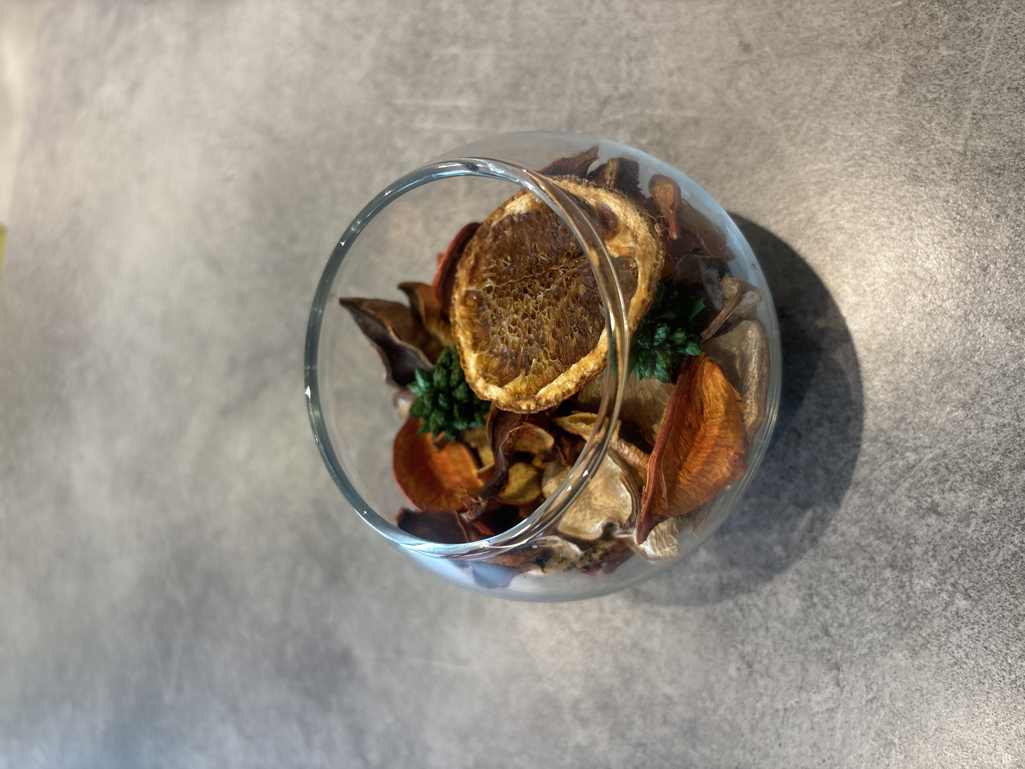 The image depicts a glass bowl containing a potpourri arrangement. The potpourri includes dried orange slices, dried flower petals in shades of brown, beige, and orange, and small green floral accents. The bowl sits on a textured gray surface. There is nothing in the image to indicate a specific location such as Burgas, Bulgaria.
