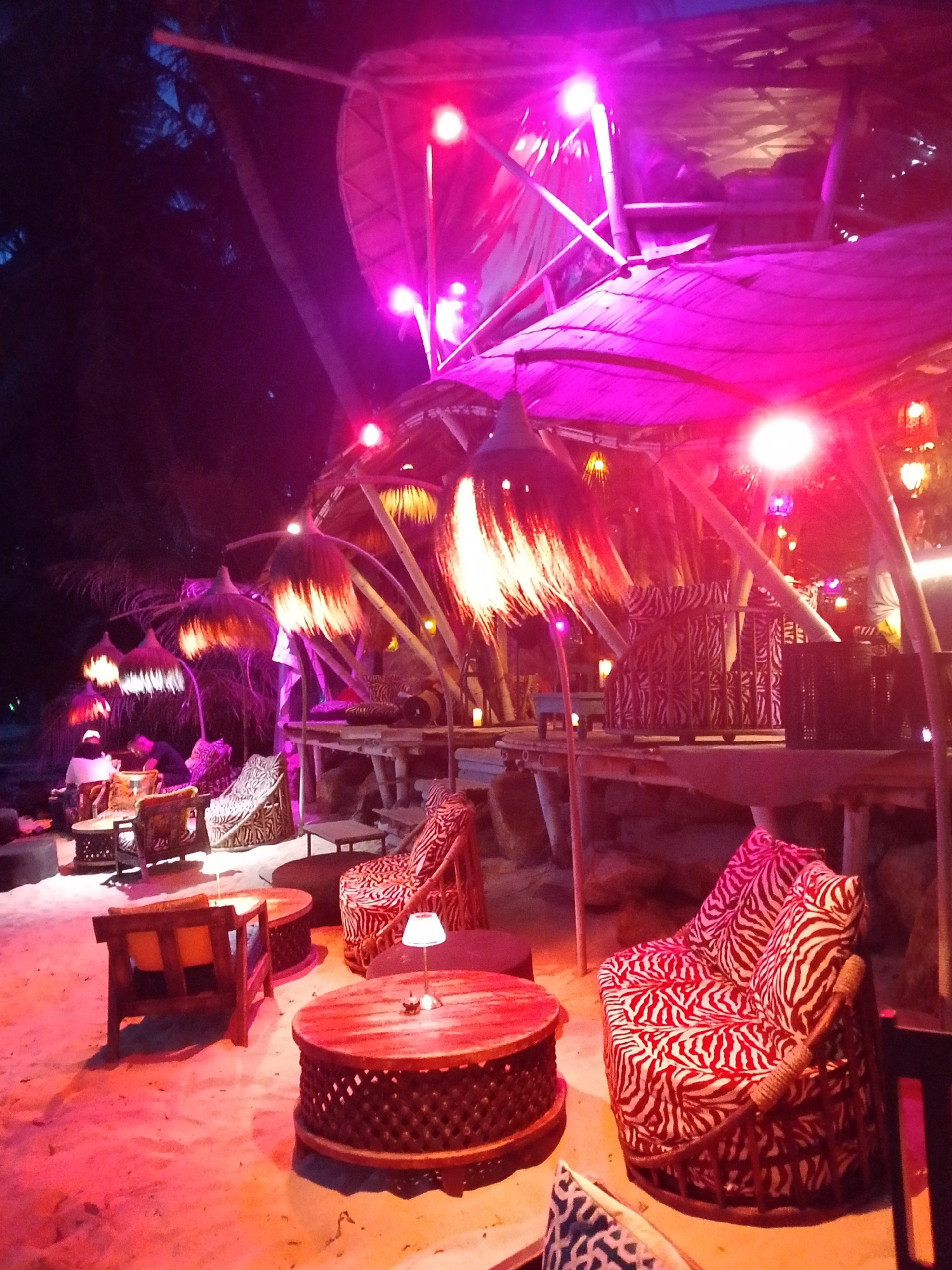 The image depicts an outdoor seating area, likely a bar or restaurant, illuminated by vibrant pink and purple lighting at night. The setting appears to be on a sandy surface, possibly a beach, with several tables and chairs arranged. In the foreground, there are two round, carved wooden tables with intricately patterned bases. A small, white-shaded lamp sits on one of these tables. To the right, a large armchair with a distinctive red and white zebra print upholstery is prominently featured. Another similar armchair is visible further back, to the left of the main one. Behind these seating areas, a multi-level structure with a thatched or bamboo roof is visible, supported by wooden beams. Several large, decorative hanging lamps with fringed shades are suspended from the structure. A few people are present, one sitting at a table in the background to the left, and another person standing near a table in the middle-ground. The overall atmosphere is tropical and festive, characteristic of a nighttime entertainment venue. The location is specified as Tambon Koh Phangan, Thailand.