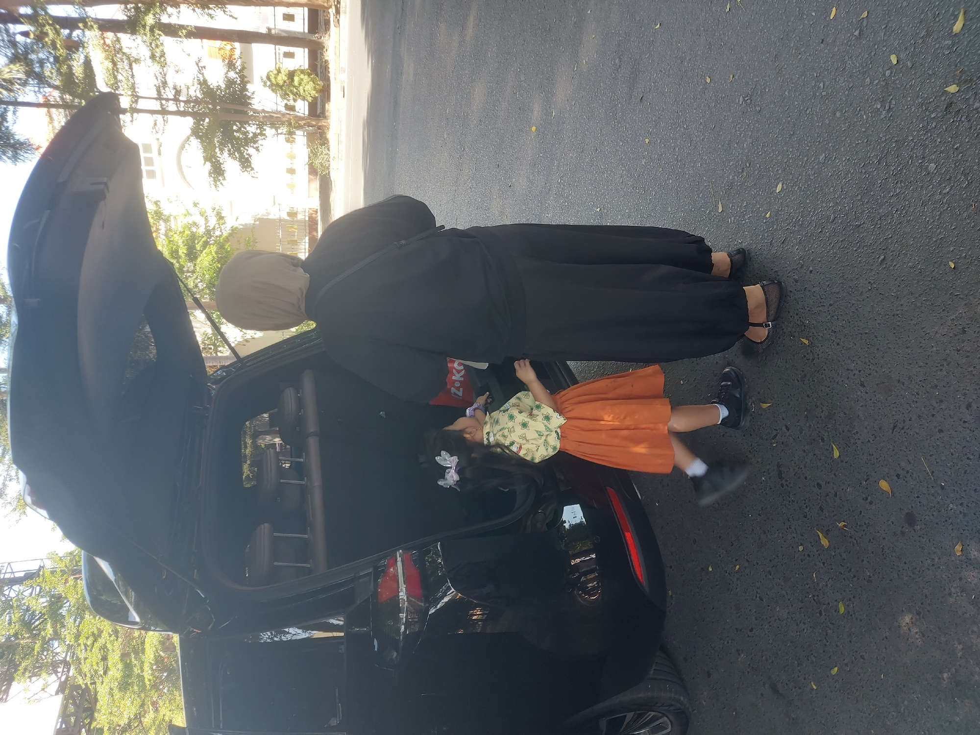A black SUV is parked on an asphalt road, with its rear hatch open. Inside the cargo area, a child is standing and looking to their right. The child wears a light-colored shirt with a pattern of green leaves and small yellow flowers, and an orange pleated skirt. They have black hair with a light pink bow on the right side.
A woman wearing a light beige hijab and a black long-sleeved top and loose black pants stands beside the child, facing away from the camera. She has a black bag slung over her right shoulder. The woman appears to be interacting with the child or looking into the cargo area.
In the background, trees with green foliage line the street, and a white building with arched windows and a tiled roof is visible. The sky is clear and bright. The scene is set during the day, likely in a residential or public area within the city of Makassar, Indonesia, suggested by the tropical trees and architectural style of the building.