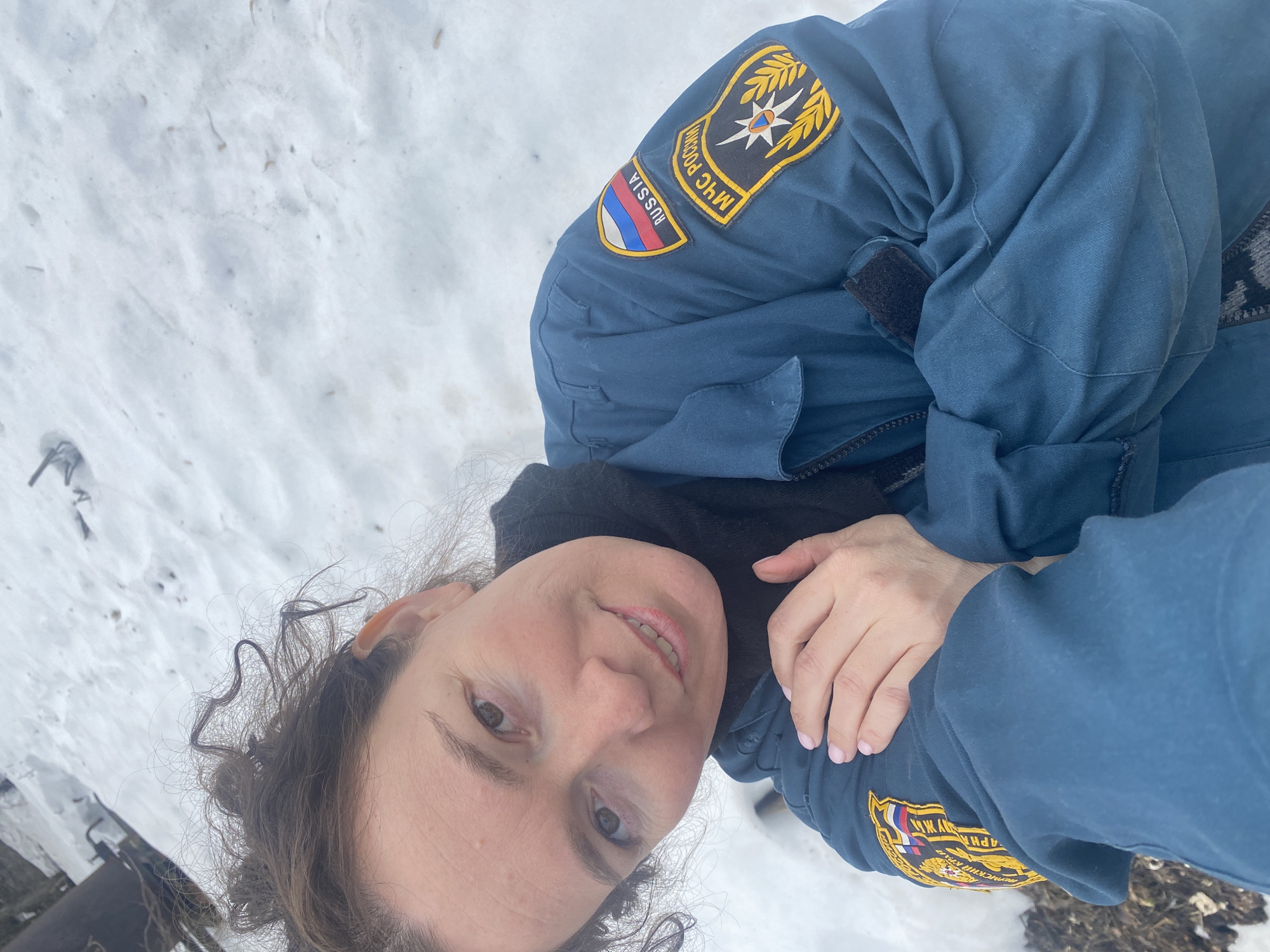 A woman lies in the snow in Ольховка, Russia. She is wearing a dark blue uniform with patches identifying her as a member of the Russian Ministry of Emergency Situations (МЧС России). Patches depict the Russian flag and the ministry's emblem. The snow is white and covers the ground. Her expression is neutral.