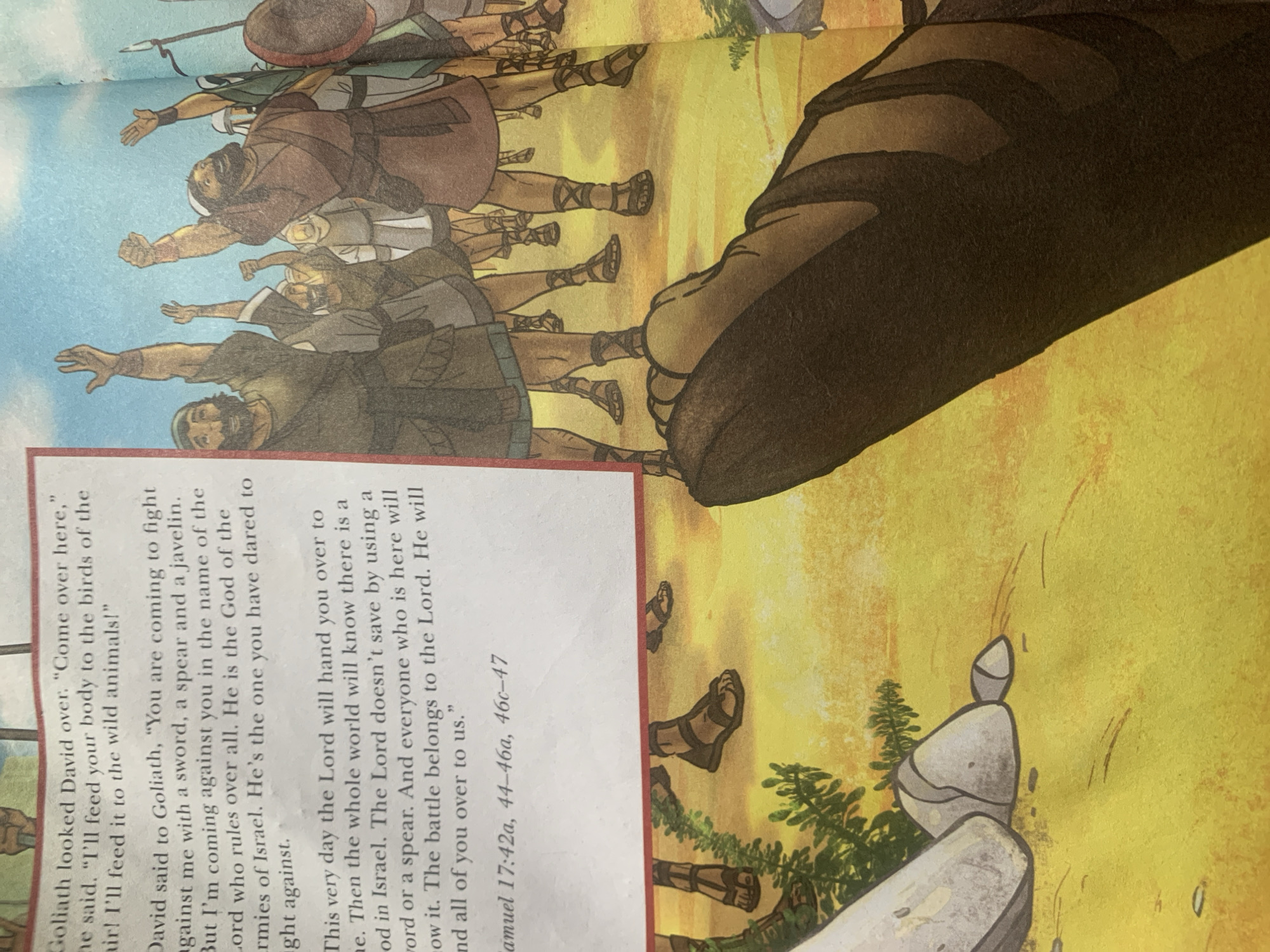 The image depicts a page from a book, with text on the left and an illustration on the right. The text is about a dialogue between Goliath and David. The illustration shows a group of people in historical attire, with their arms raised in the air, possibly cheering or in a celebratory mood. In the foreground of the illustration, the lower legs and feet of a person wearing sandals are visible, suggesting a close-up perspective. The background of the illustration is a bright yellow, possibly representing a sandy or sunny environment. There are rocks and some greenery in the lower left portion of the illustration. The provided information about the location of Jauro Yinu, Nigeria, is not directly supported by the visual content of the image.