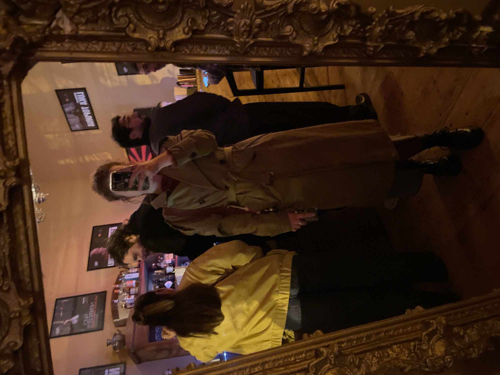 The image shows a reflection in an ornate, gold-colored mirror. The reflection depicts a group of people in what appears to be a bar or pub. One person is holding up a smartphone, seemingly taking a selfie. The people appear to be casually dressed. The background includes shelves stocked with bottles, likely liquor. Framed posters or prints are visible on the wall. The location is identified as Tbilisi, Georgia.