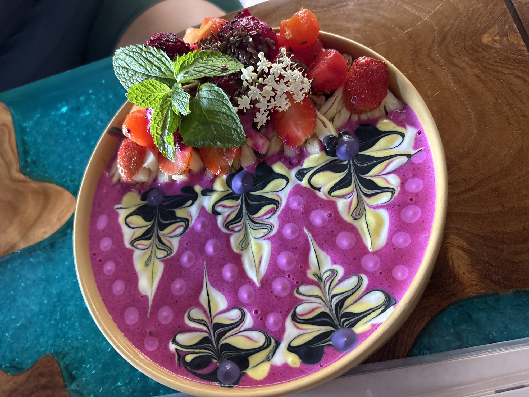 A high-angle, close-up shot presents a vibrant smoothie bowl, likely photographed in the Ubud District of Indonesia, judging by the tropical-style presentation. The bowl, with a light-colored rim, is filled with a bright pink smoothie base that is decorated with artistic swirls of white, yellow, and black patterns reminiscent of stylized leaves or flowers. Round pink decorations are spaced uniformly across the smoothie. The bowl is topped with an assortment of fresh fruits, including sliced strawberries, diced tropical fruit (possibly dragon fruit and/or papaya), blueberries, and what appears to be sliced banana. Fresh mint leaves and small white flowers provide an additional garnish. The bowl is placed on a wooden surface with a blue resin swirl resembling water. Part of a person's arm is seen at the top of the picture, suggesting the photo was taken by someone holding the bowl.