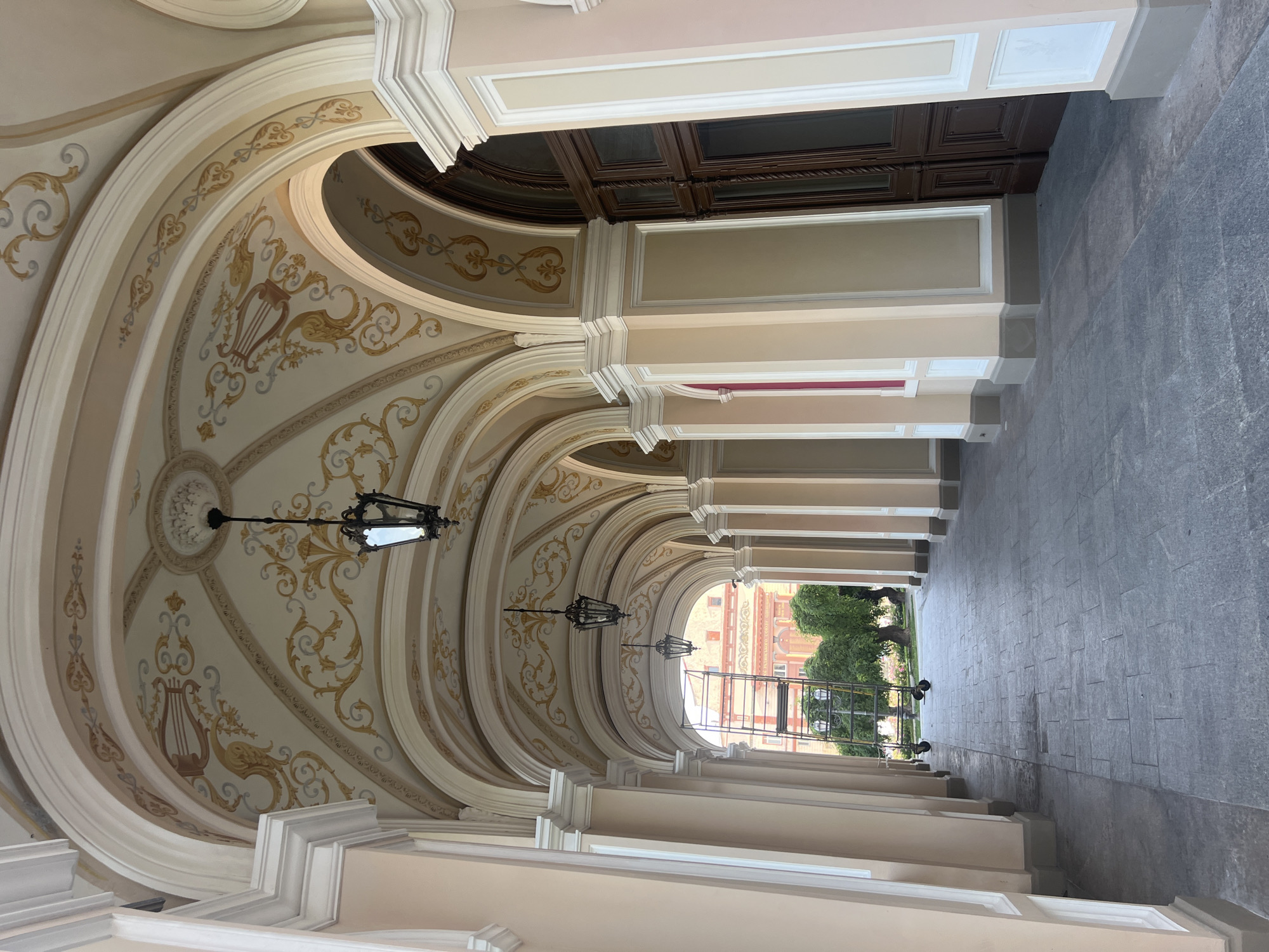 The image depicts a wide, arched portico with ornate ceiling frescoes and hanging lanterns. The portico is lined with columns and has large wooden doors on the right side. The floor is paved with dark gray stone tiles. In the background, through the arches, a building under renovation with scaffolding is visible, along with trees and street lamps. This scene is located in the city of Odesa, Ukraine, specifically at the Potemkin Stairs.