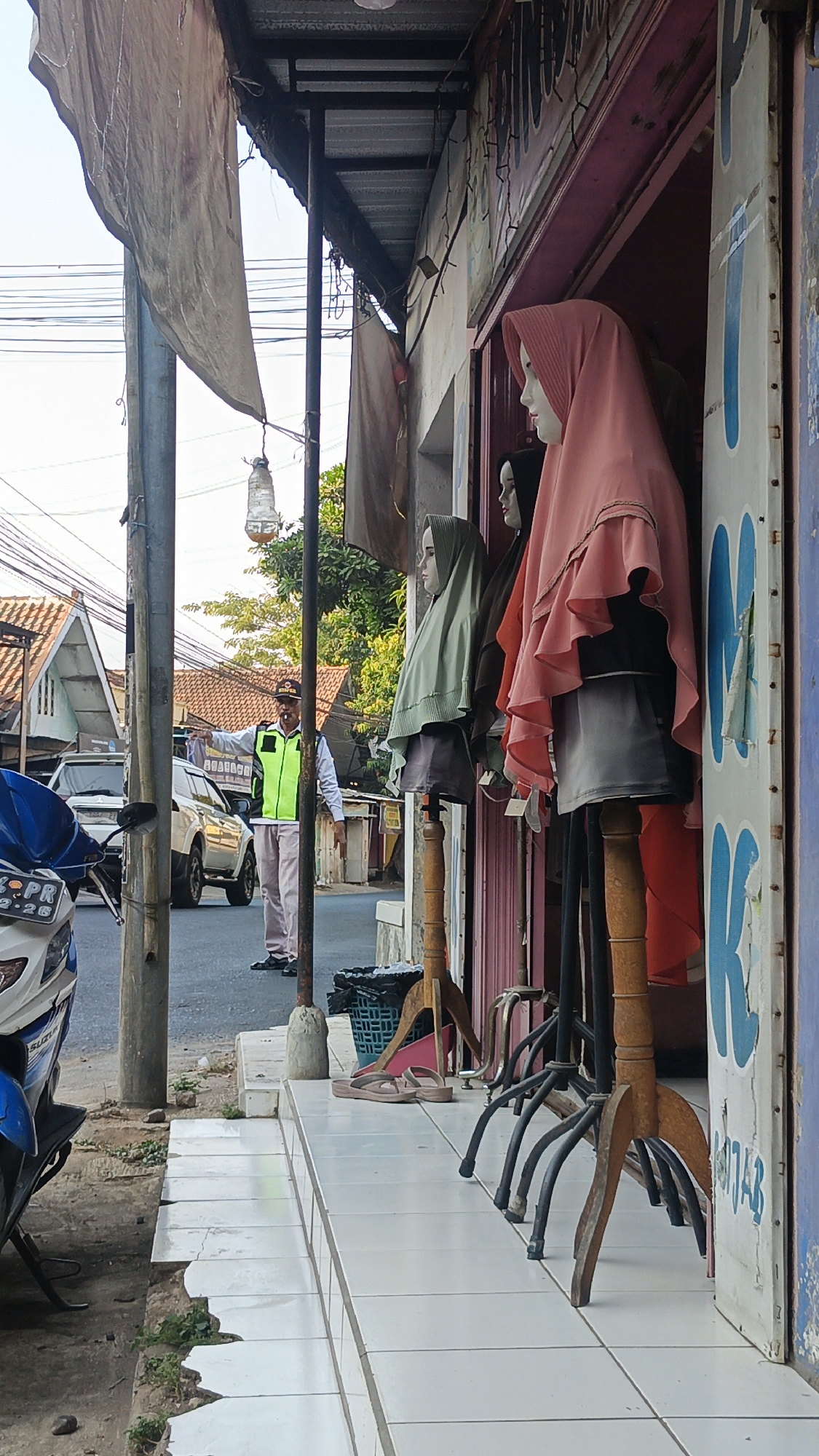 In Dawe, Indonesia, a man in a high-visibility vest directs traffic on a street. In the foreground, mannequins are displayed outside a shop, dressed in hijabs. The scene captures a slice of urban life with elements of commerce and public order.