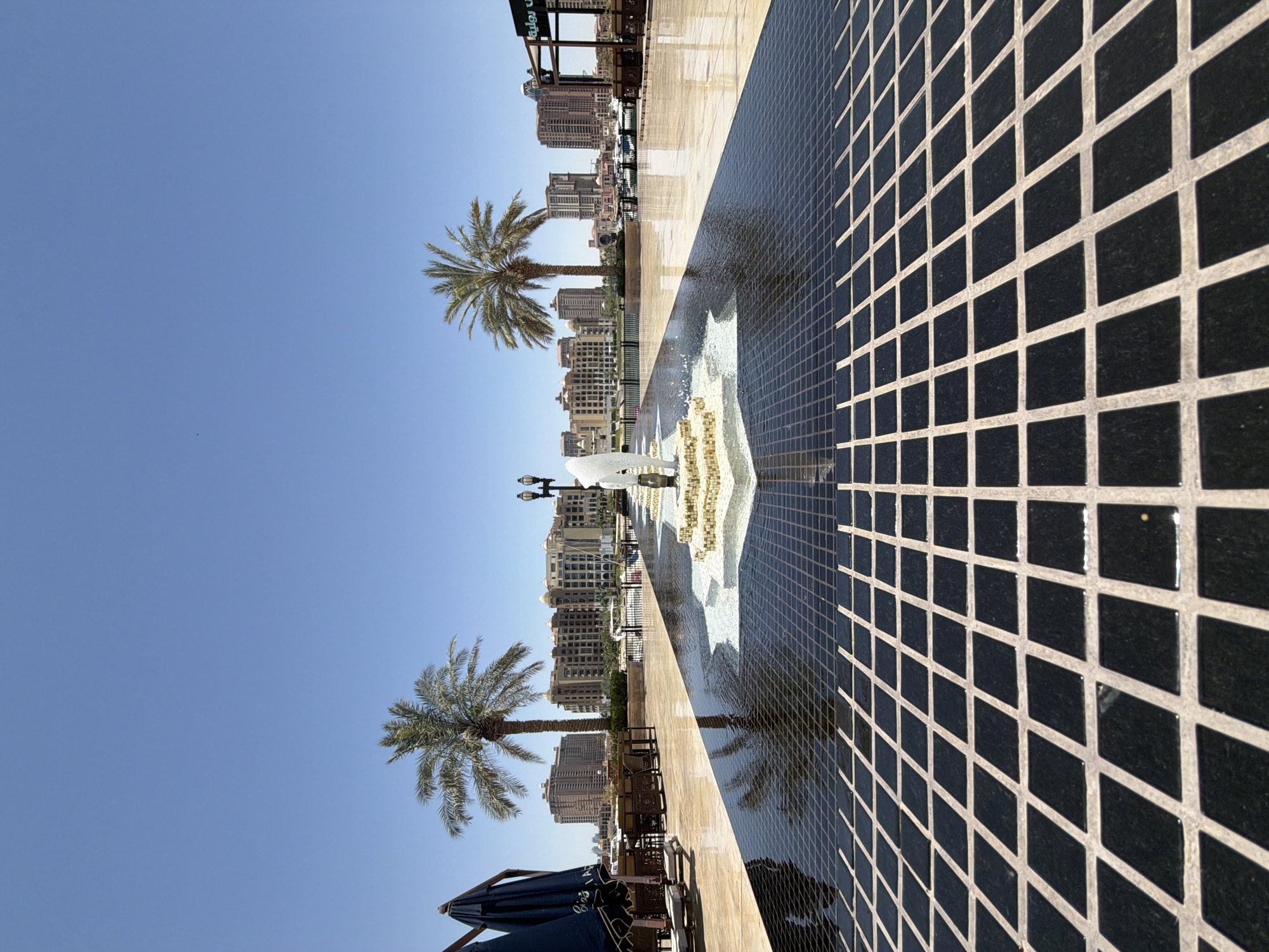 A wide-angle outdoor shot shows a reflective water fountain area in front of a cityscape. The foreground is dominated by a rectangular pool with a dark, tiled surface featuring a grid pattern of square black tiles separated by light grout. Water from the fountain creates ripples on the surface of the pool. The fountain itself is in the center of the image, with multiple streams of water spraying upwards from a multi-tiered, ornate structure. Beyond the fountain, the ground transitions to a polished, light-colored stone surface. Two palm trees are visible in the mid-ground, with one taller, more mature tree on the left and another slightly shorter one on the right. Behind the trees, a row of multi-story buildings with arched windows and various architectural styles forms the cityscape. The sky is clear and blue, indicating a sunny day. To the far left, there are several dark blue umbrellas and tables, suggesting an outdoor seating area for a cafe or restaurant. In the far background, there are boats docked in a body of water. The overall scene depicts a modern urban waterfront area with a focus on water features and outdoor leisure spaces.