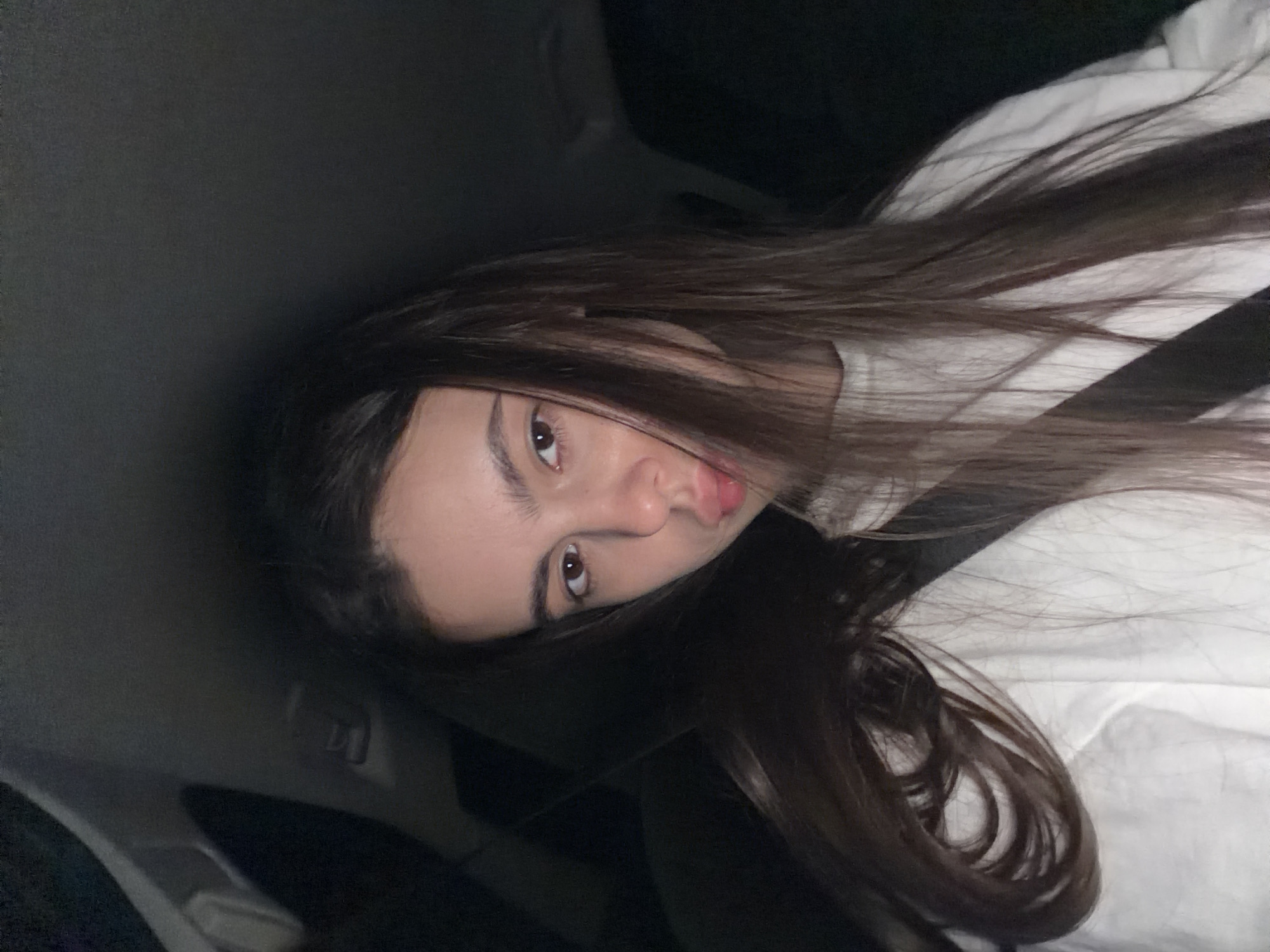 A young woman with long, dark brown hair and brown eyes is pictured from the chest up, looking at the camera. She is wearing a white crewneck sweatshirt and a black seatbelt across her chest. Her lips are pursed. The background appears to be the interior of a car at night, with dark upholstery and what looks like the headliner of the car visible. There are no discernible landmarks or details that would indicate a specific location within the city of Chaikovsky, Russia.