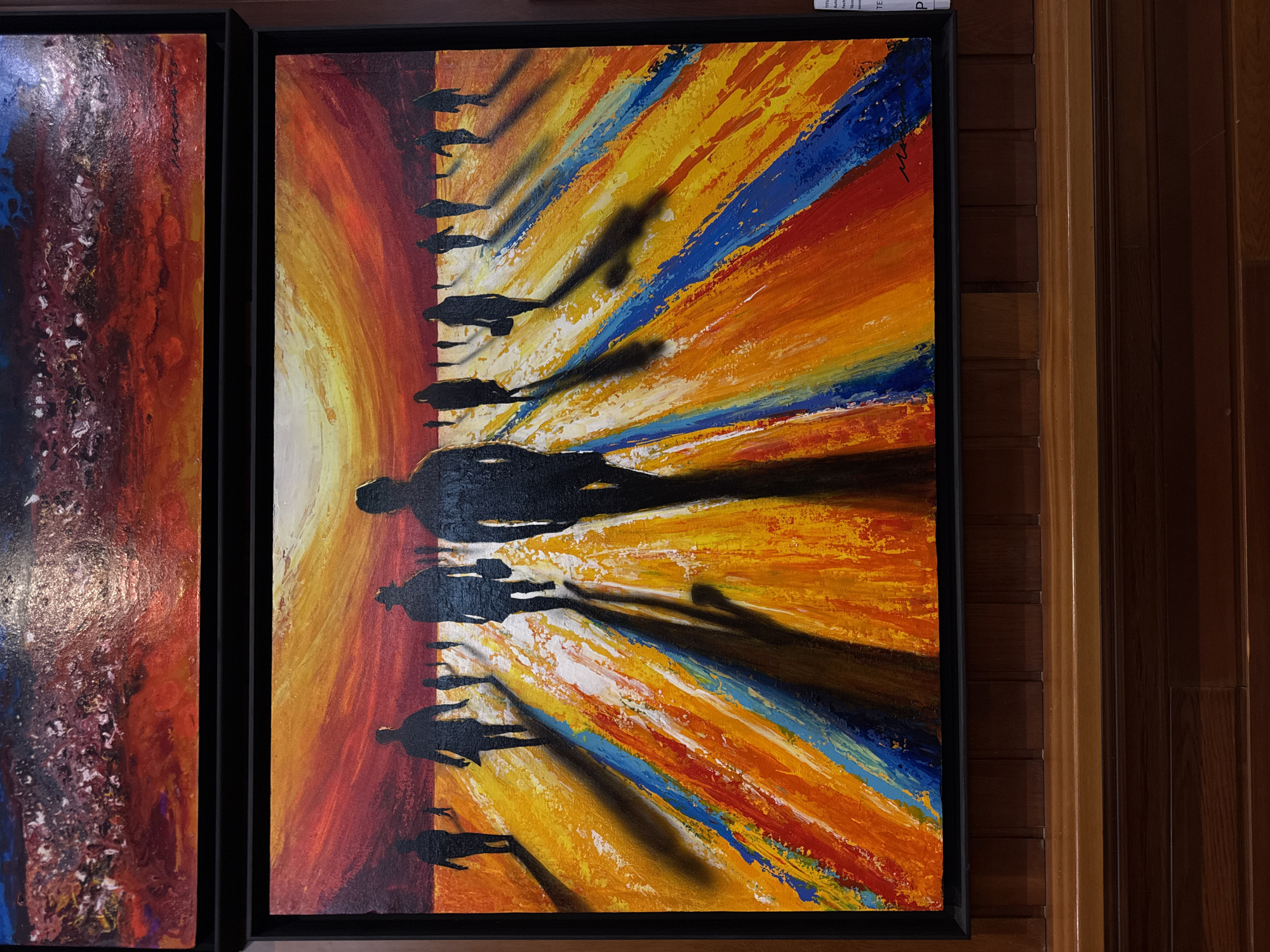 The image displays two framed paintings hanging on a dark wooden wall. The top painting is partially visible and appears to be an abstract depiction of a turbulent, reddish-purple sky. The larger, bottom painting features a group of silhouetted figures walking away from the viewer towards a vibrant, sunset-like sky that transitions from fiery red to yellow. The ground is rendered with thick, textured strokes of orange, yellow, blue, and white, creating a sense of depth and movement. Long, dark shadows stretch out from each figure, contributing to the dramatic atmosphere. The overall style is expressive and colorful, with a focus on conveying emotion through bold brushwork and contrasting hues. A small white label is attached to the lower right corner of the bottom painting's frame, though its text is largely illegible. The paintings are displayed in a well-lit indoor environment, likely an art gallery or exhibition space. There is no indication in the image that this scene is located in Torrejón de Ardoz, Spain.